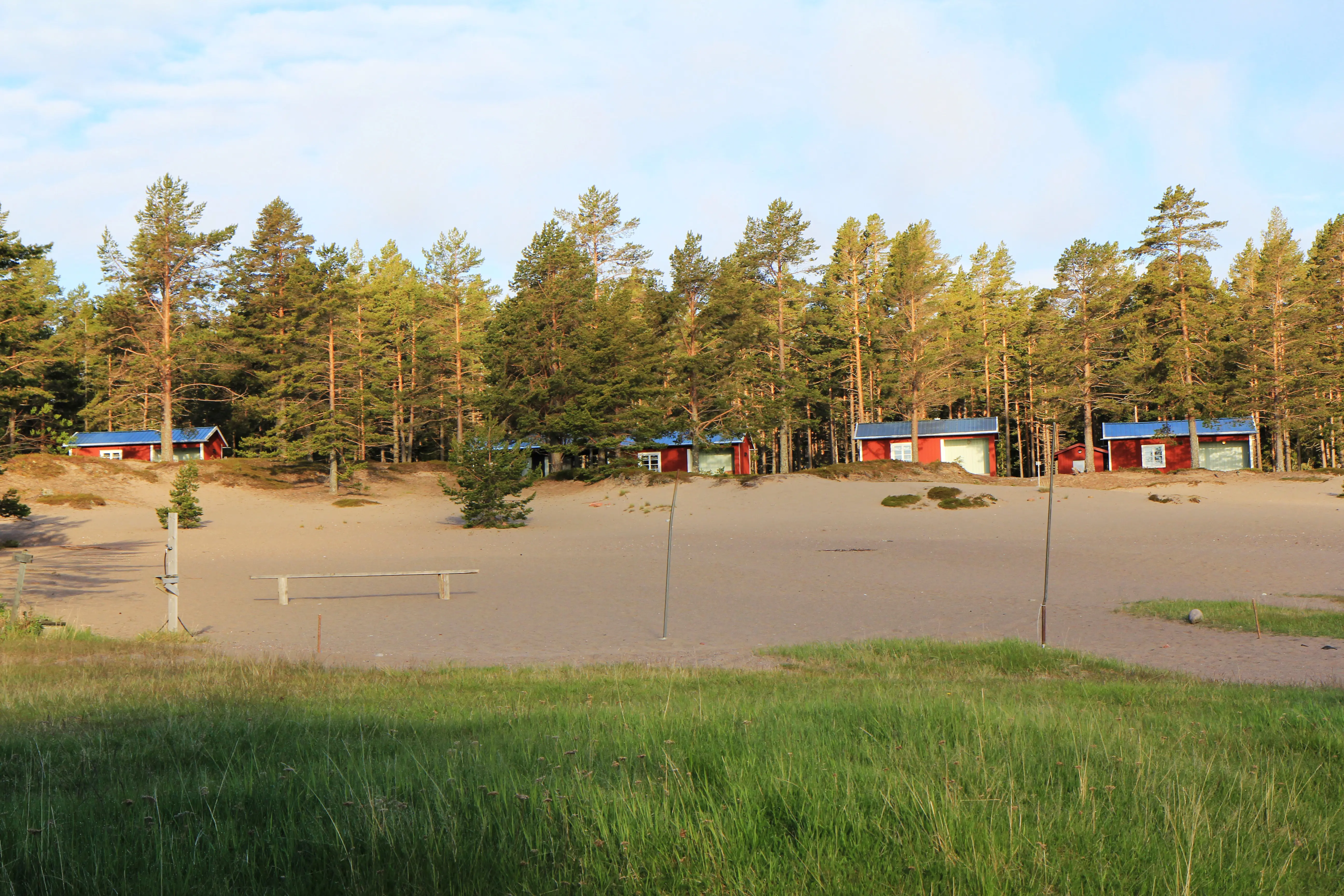 Nyare, röda stugor vid Sandvikens fiskeläge. Bakom dem växer tallar, framför dem är fin sand och gräs.