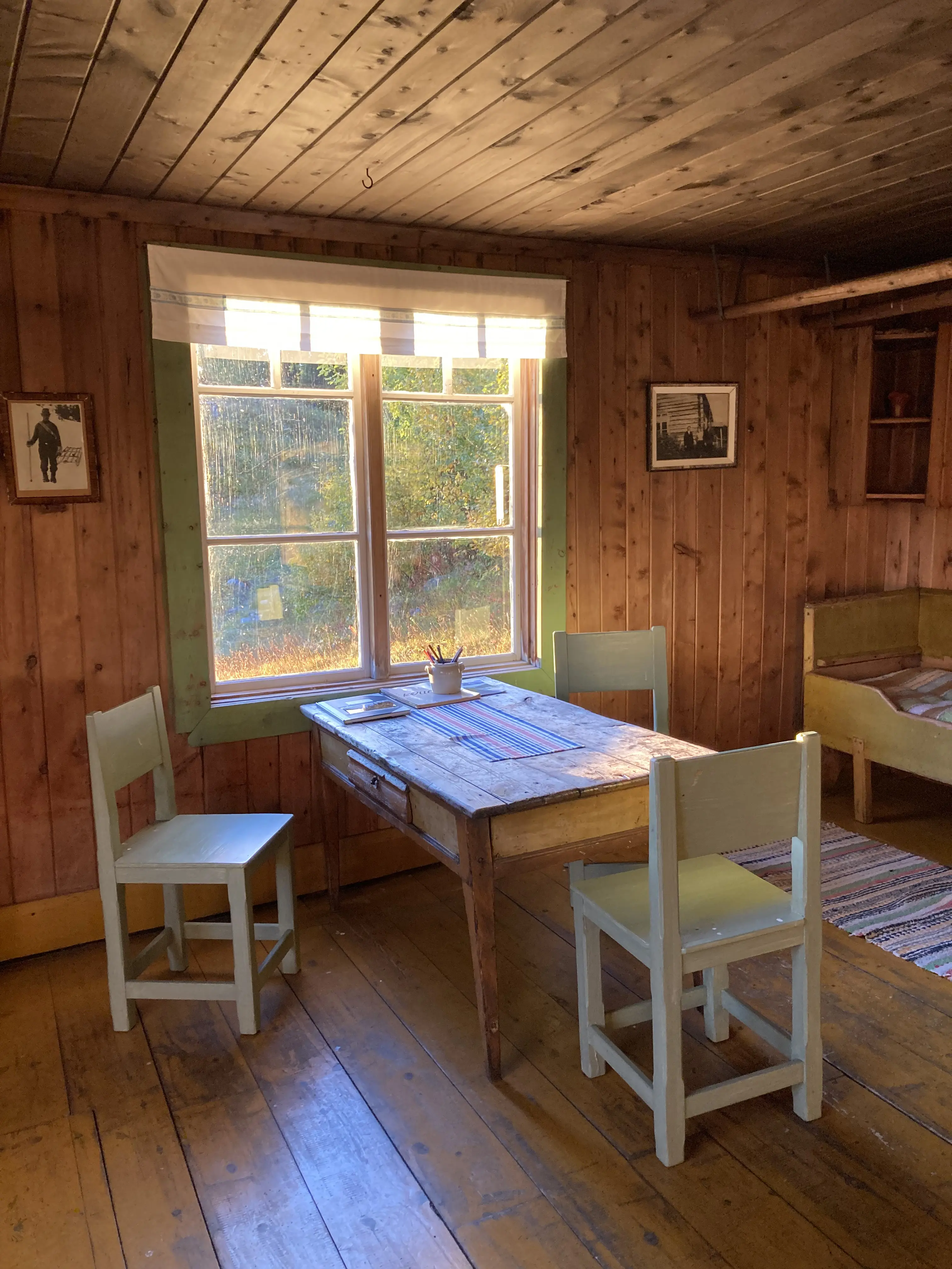 Örnbo fjäll-lägenhet, interiör. Solen lyser in genom fönstret i en stuga. Framför fönstret står ett enkelt träbord och tre trästolar i en grågrön nyans.