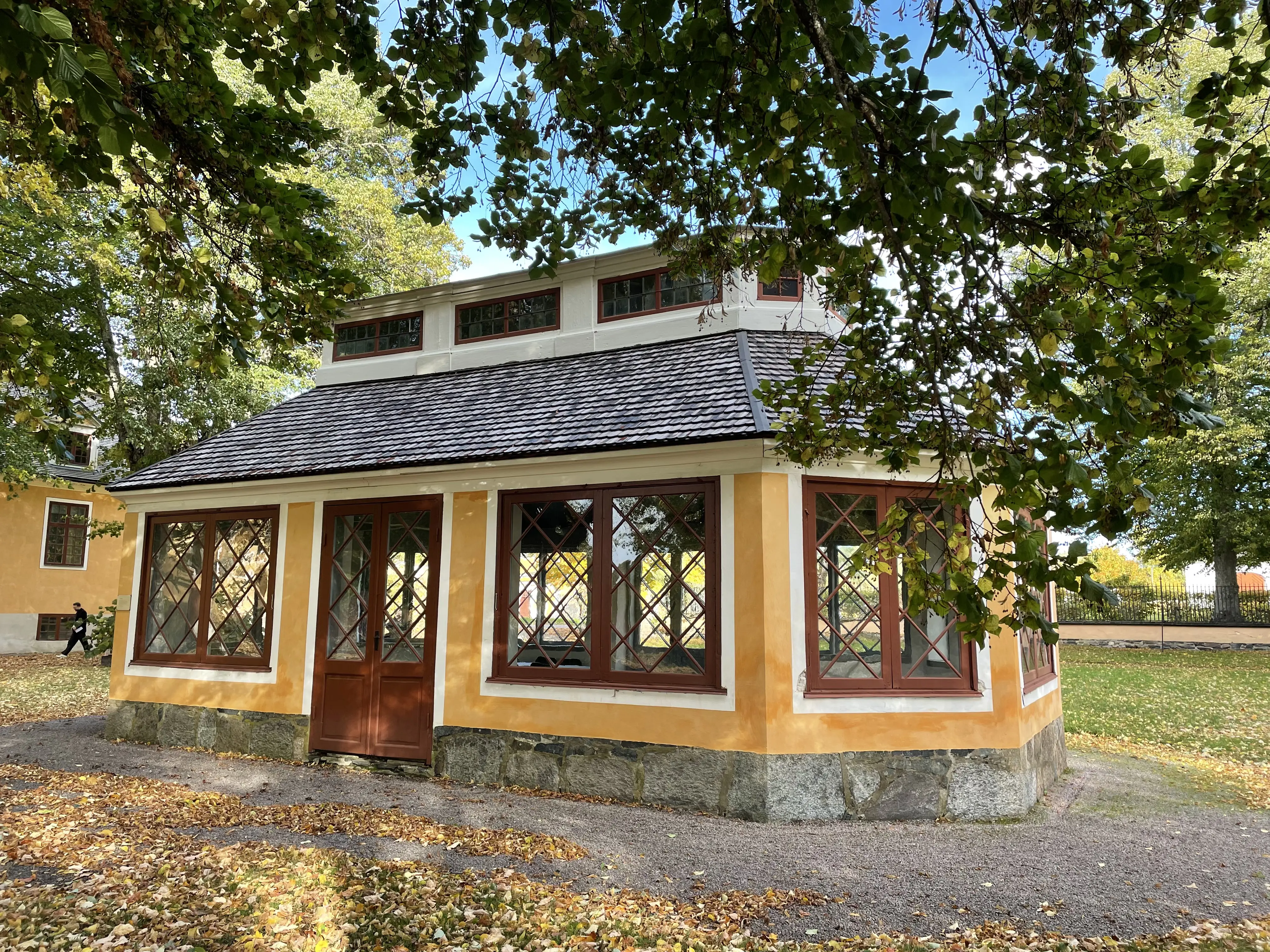 Volieren i Lövstabruks park. En liten gulputsad stenbyggnad med stora, spröjsade fönster och extra ljusinsläpp i form av en långsträckt lanternin på taket.