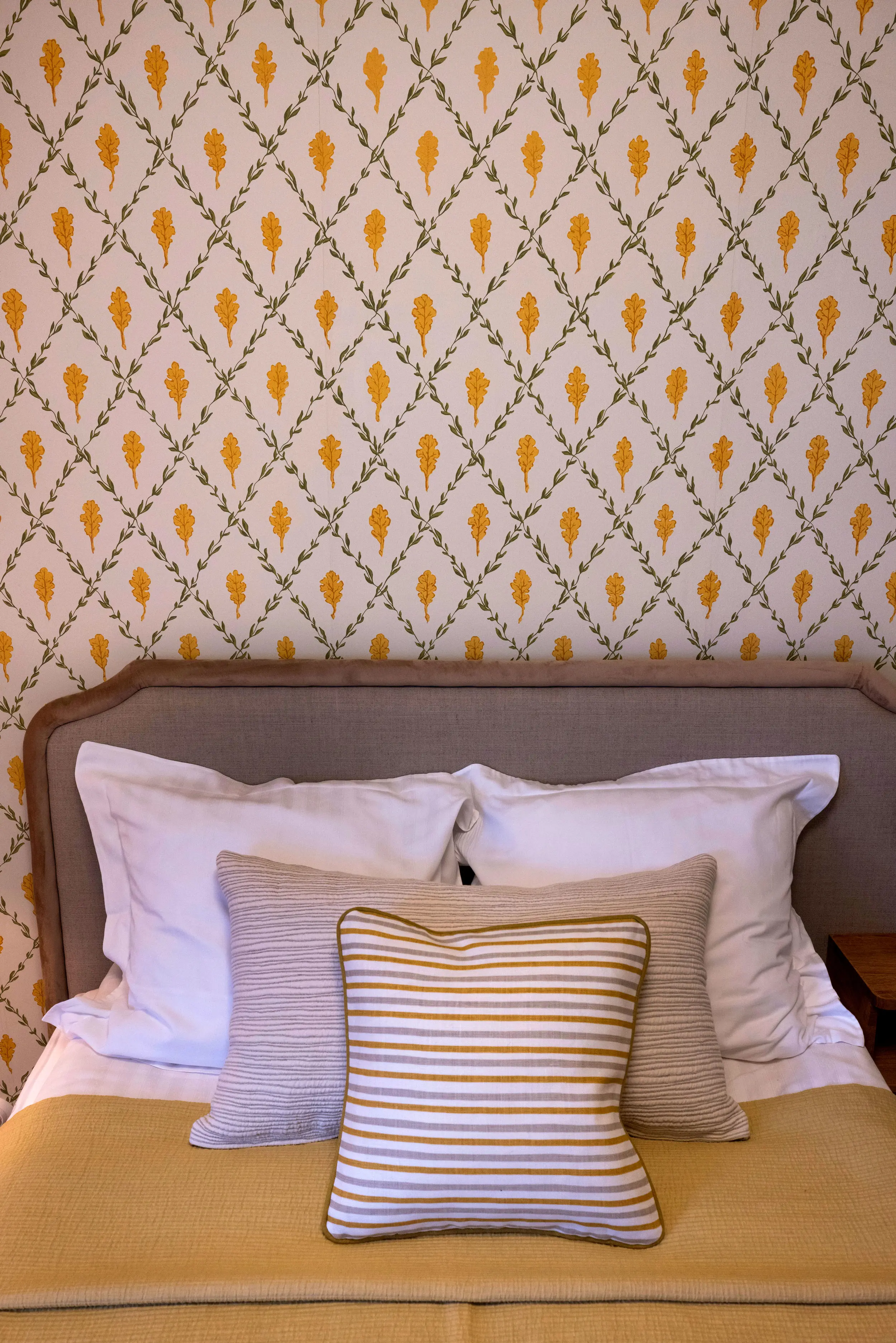 Säng framför vägg med mönstrad tapet med gula eklöv och gröna blomstergirlanger.