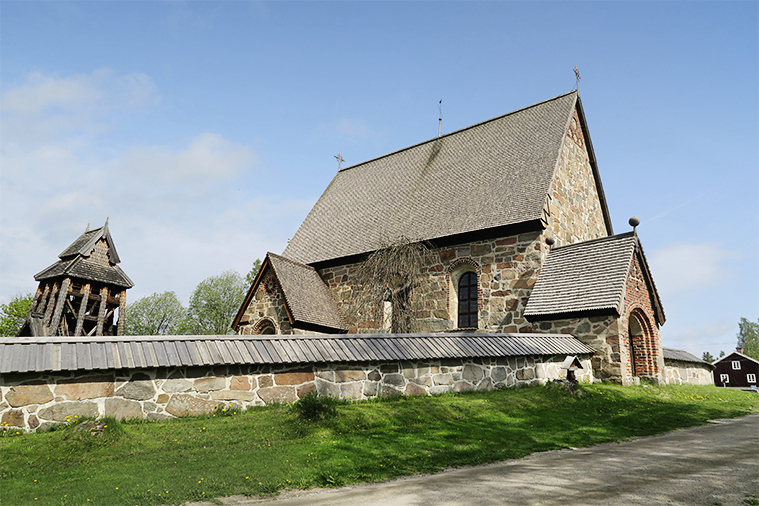 En murad kyrka med gråbrunt tak och en klockstapel i trä omges av en stenmur.