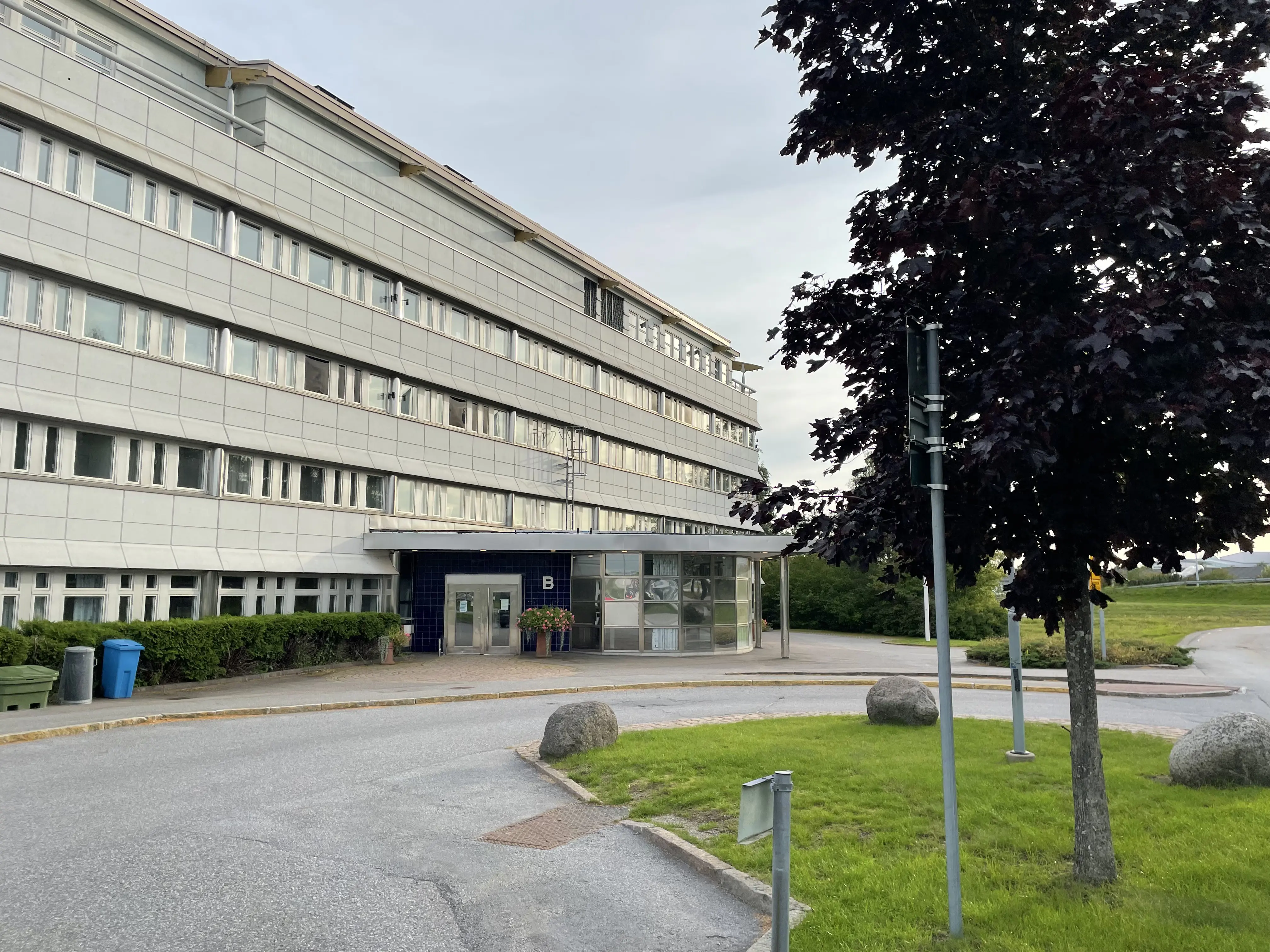 Exteriör med byggnadens framsida som vetter mot väg med lite grönska och träd i förgrunden.