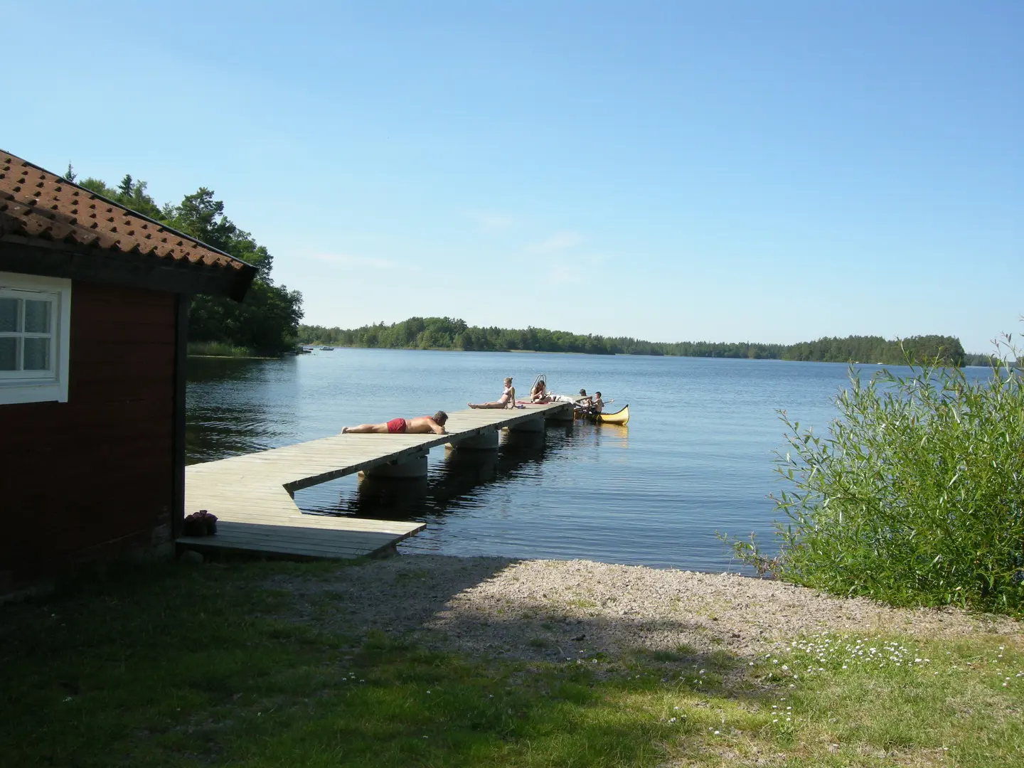 Röd stuga och badbrygga vid en strand.
