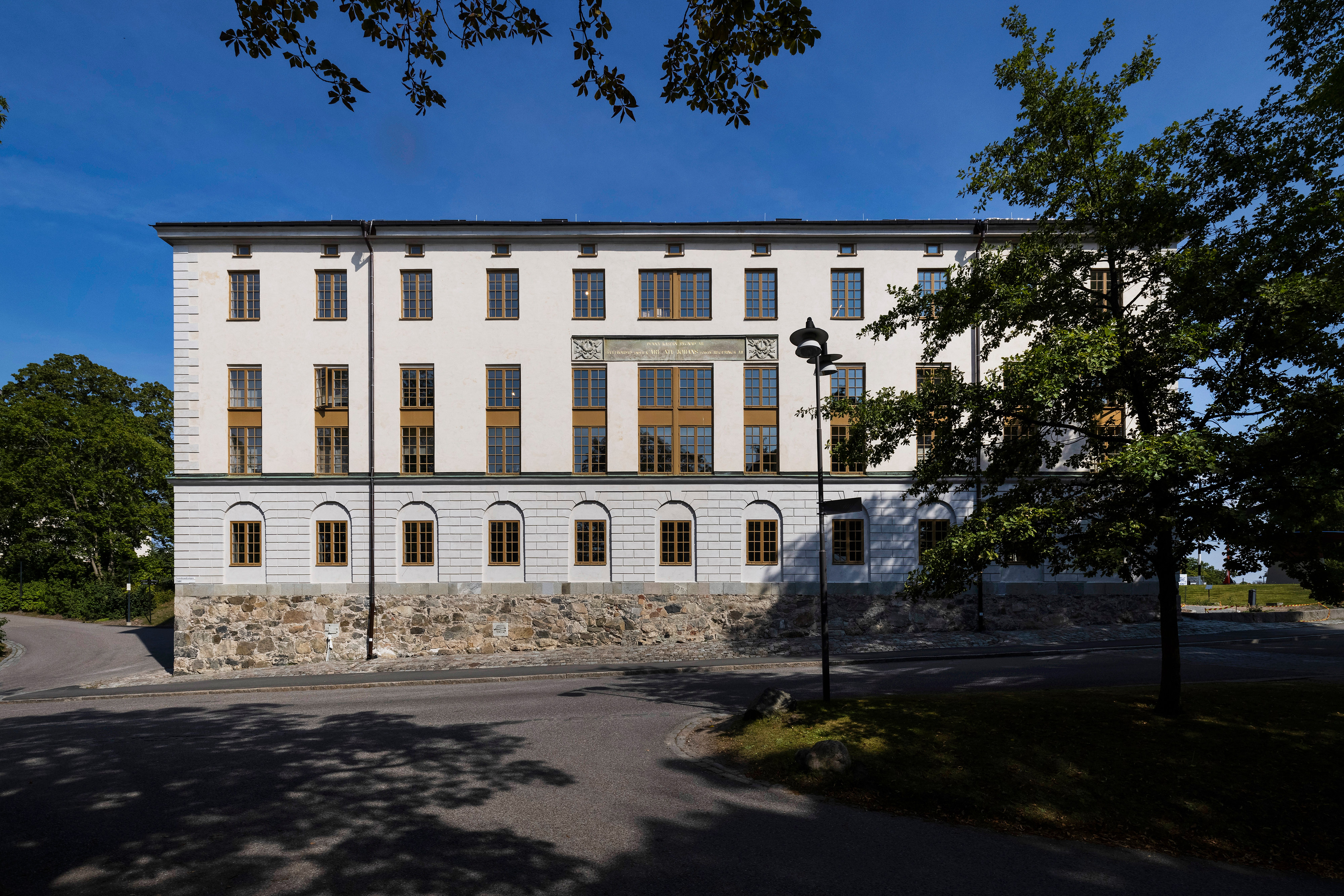 Kasern II sett utifrån vägen mot blå himmel och träd i förgrunden.