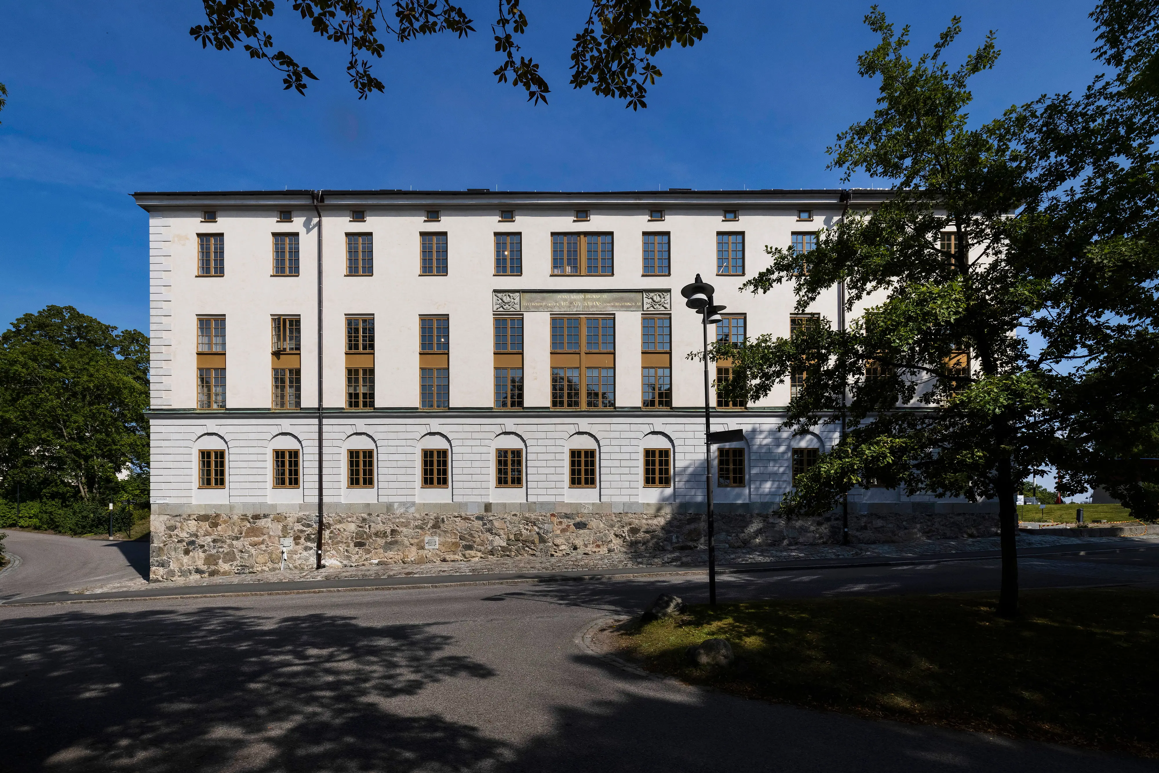 Kasern II sett utifrån vägen mot blå himmel och träd i förgrunden.