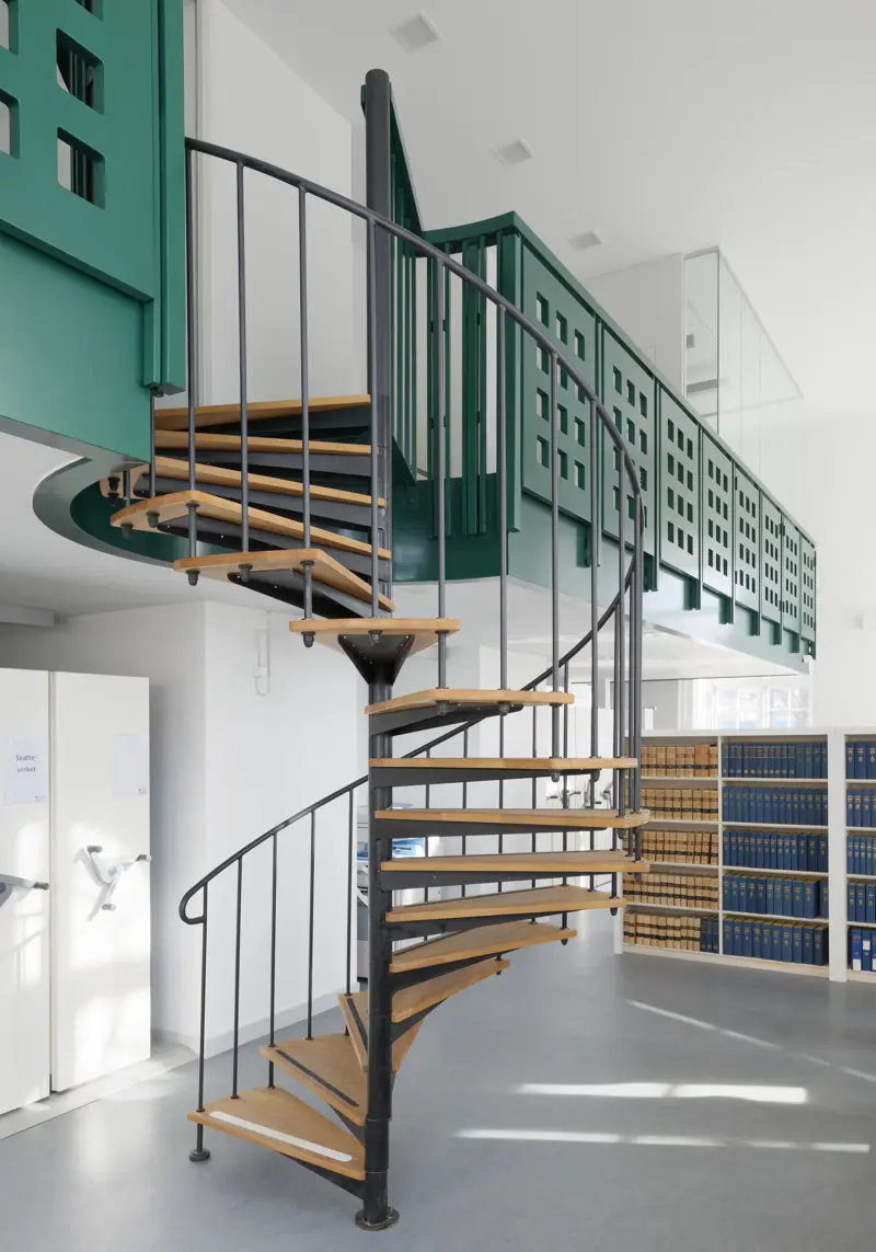 Spiraltrappa med steg av trä från en avsats med gröna räcken. Trappan går ned till ett bibliotek med bokhyllor.