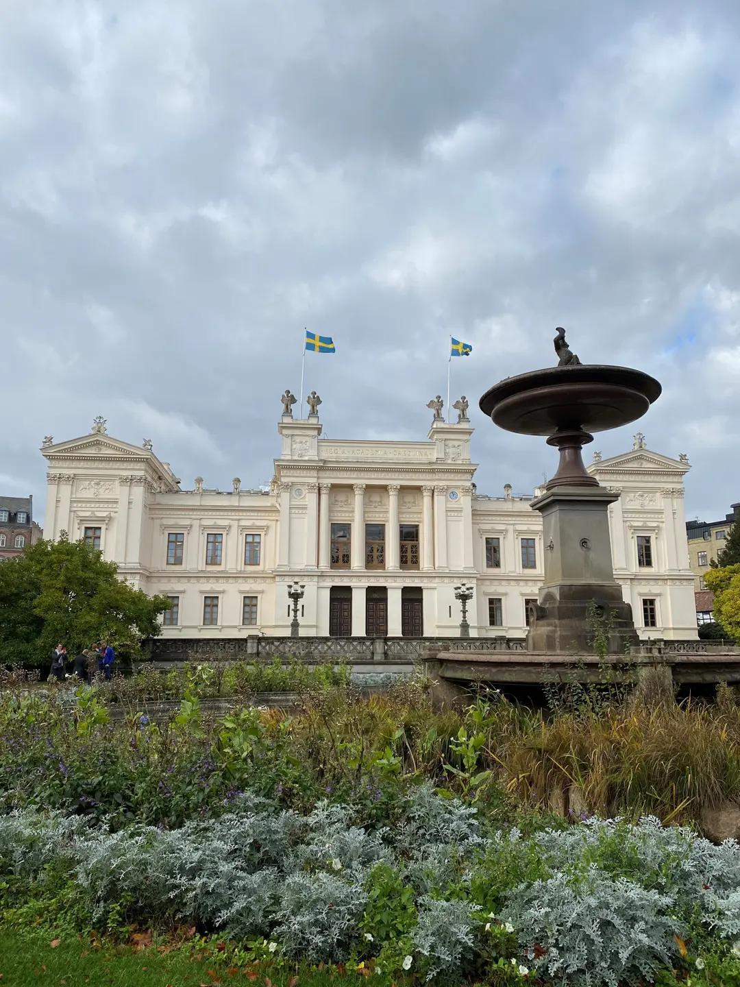 Universitetshuset sett utifrån, med en rabatt i förgrunden. På taket syns de fyra sfinxerna och två flaggstänger med Sveriges flagga.