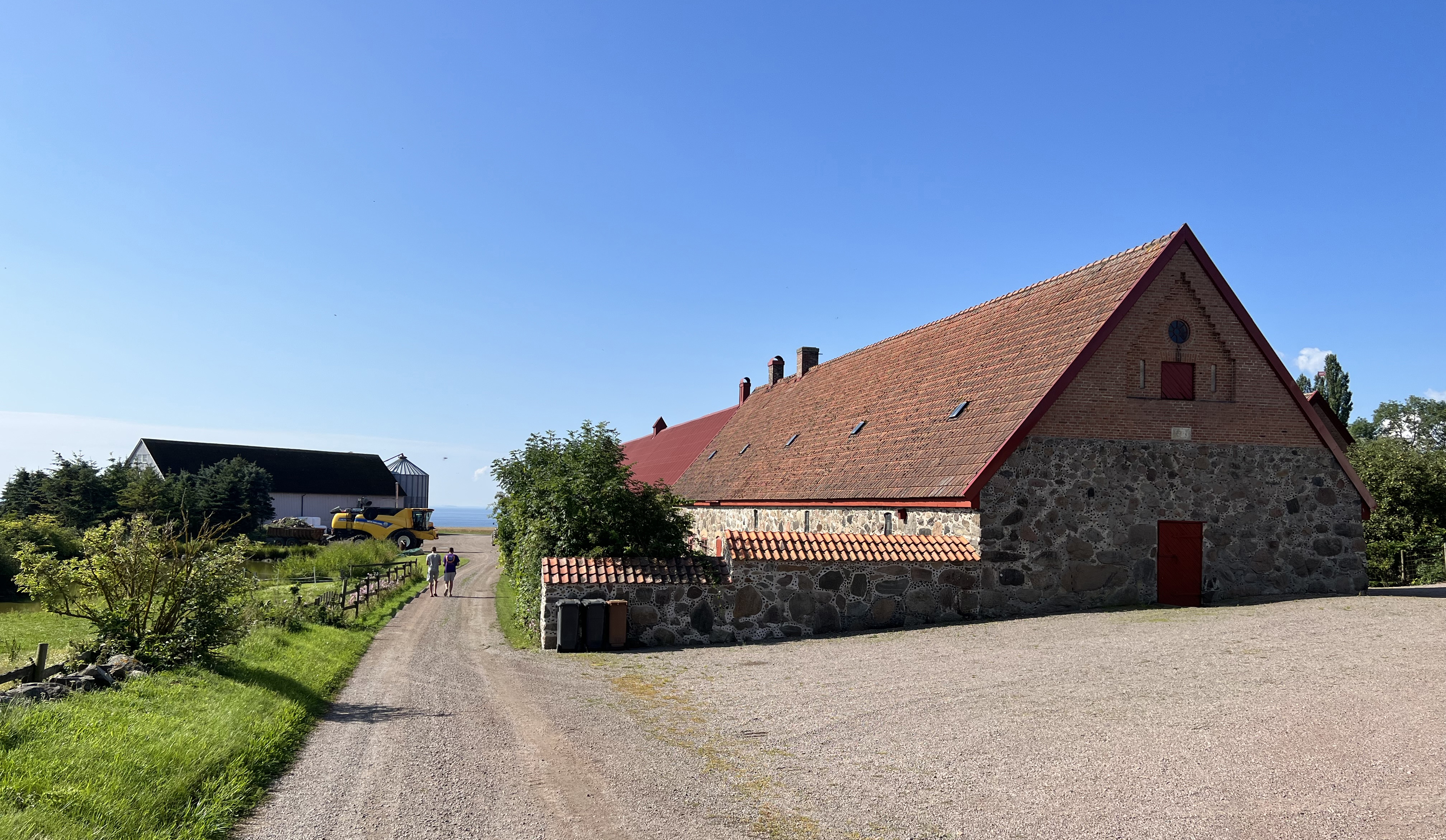 Uranienborg. En grusväg leder mellan äldre och modernare ekonomibyggnader. I fjärran skymtar havet. Sommar och grönska.