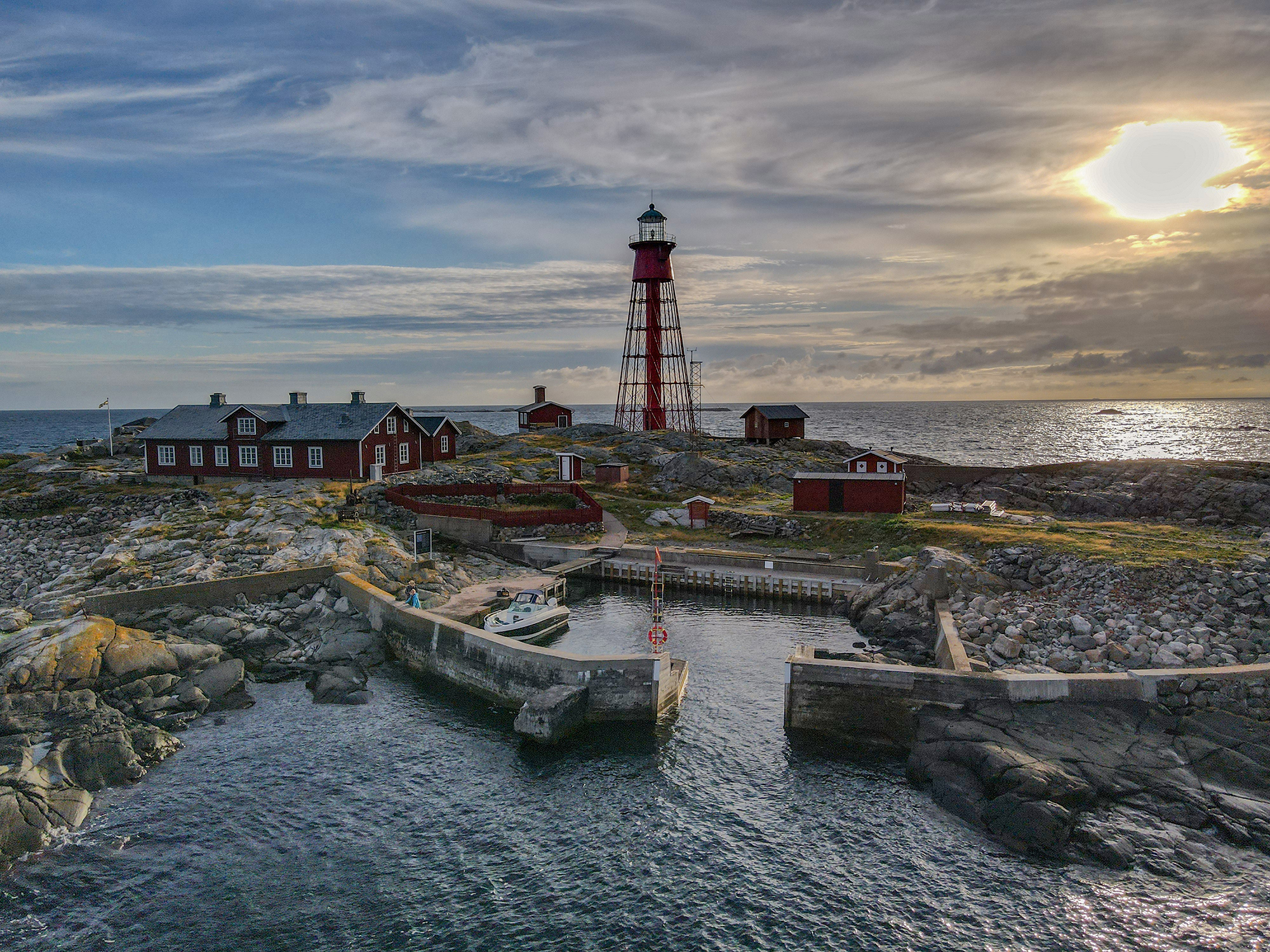 Hamneskär. Hamnen omgärdad av klippor med fyren och byggnaderna bakom. Vy mot kvällshimlen och öppet hav.