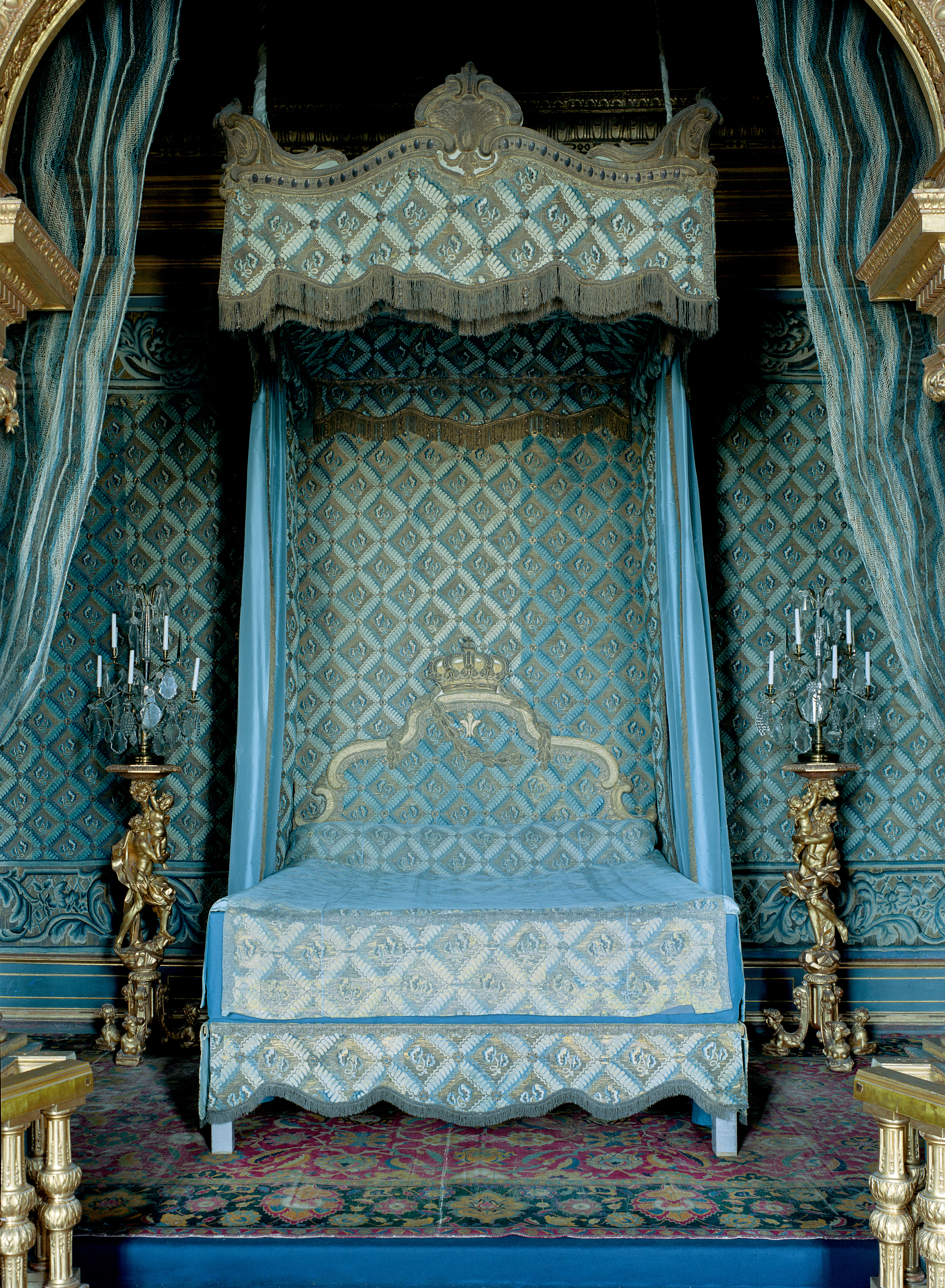Parad-sängkammaren i Drottningholms slott. I en alkov står en säng klädd i blått siden. Säng-gaveln pryds av en en förgylld krona.