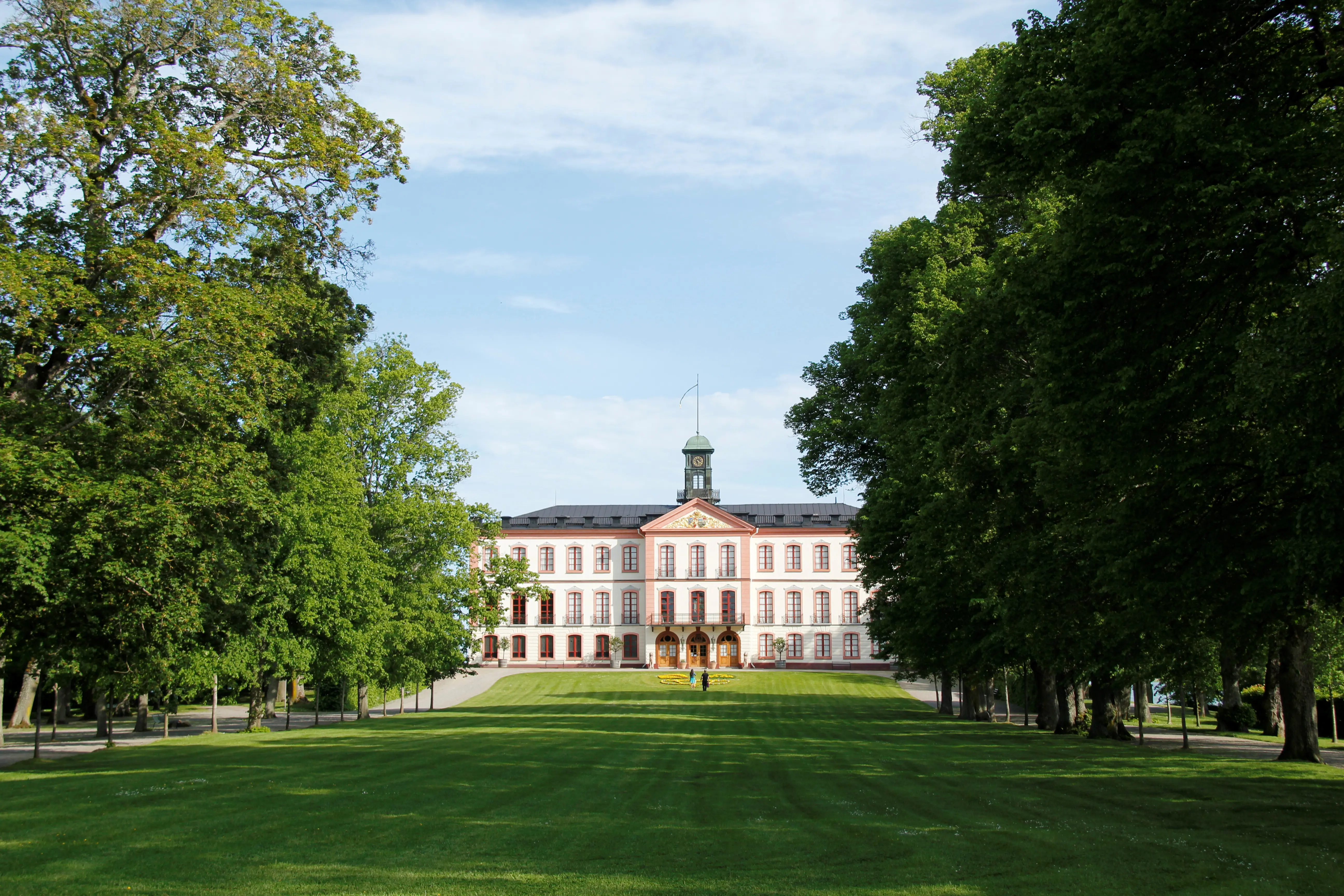 Tullgarns slott. Vitputsad byggnad i tre våningar, med röd dekor runt fönster och dörrar. På taket står ett litet klocktorn. Sommar och grönska.