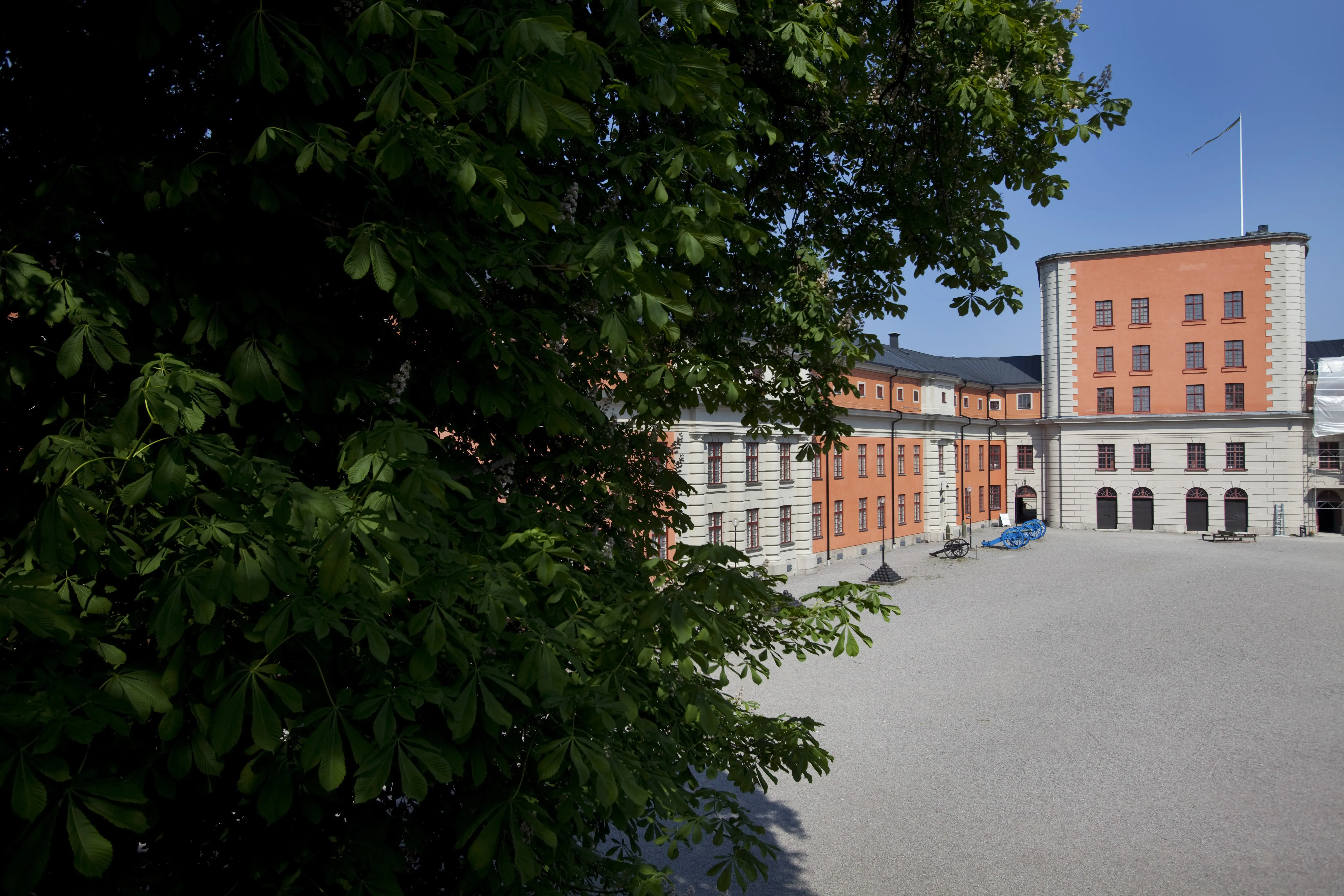 Vaxholms kastell. Innergården omges byggnader med vit och röd puts. På gården växer några stora träd. Sommar och grönska.
