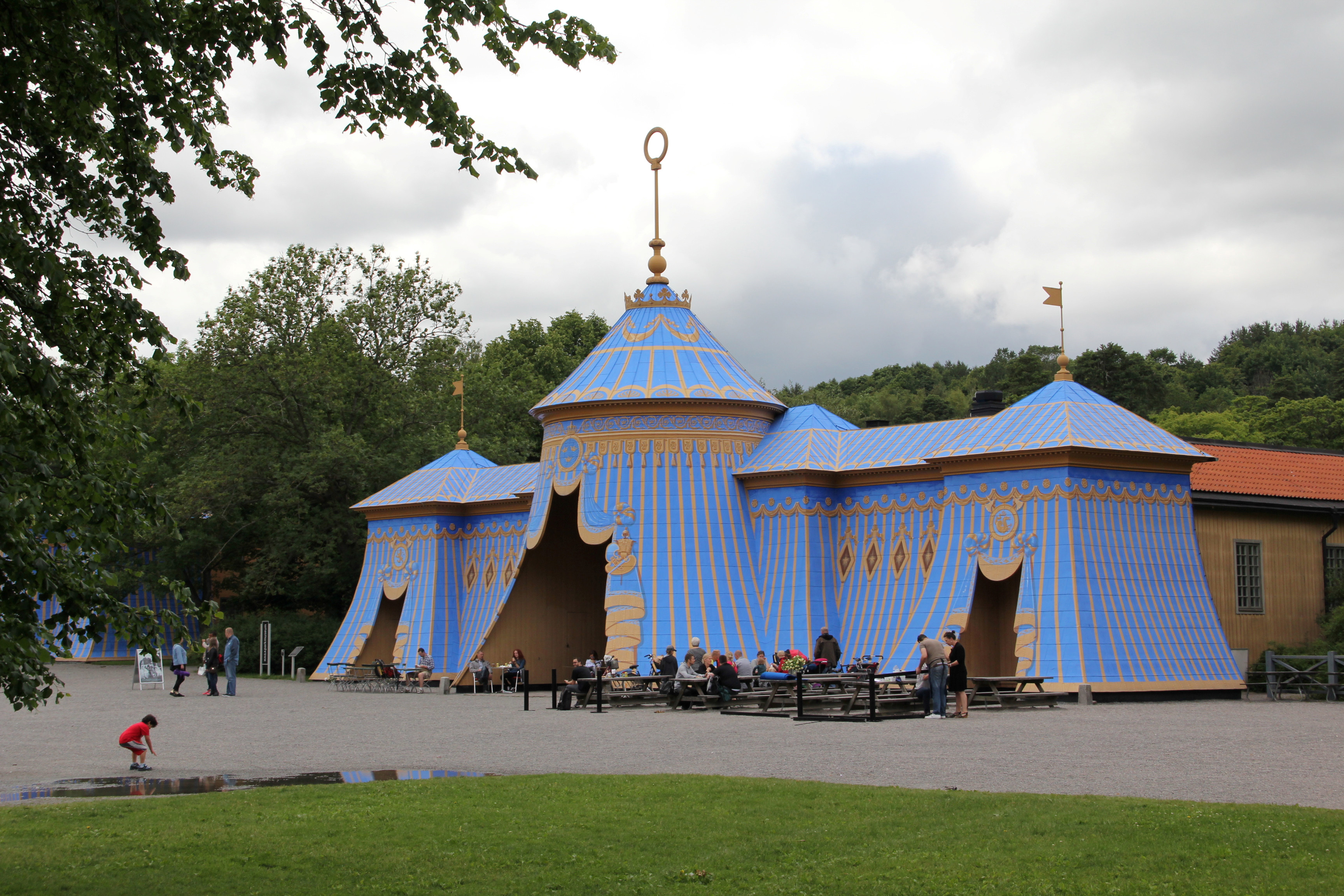 Mellersta koppartältet i Hagaparken. Besökare sitter vid bänkbord på grusplanen framför byggnaden. Sommar och grönska.