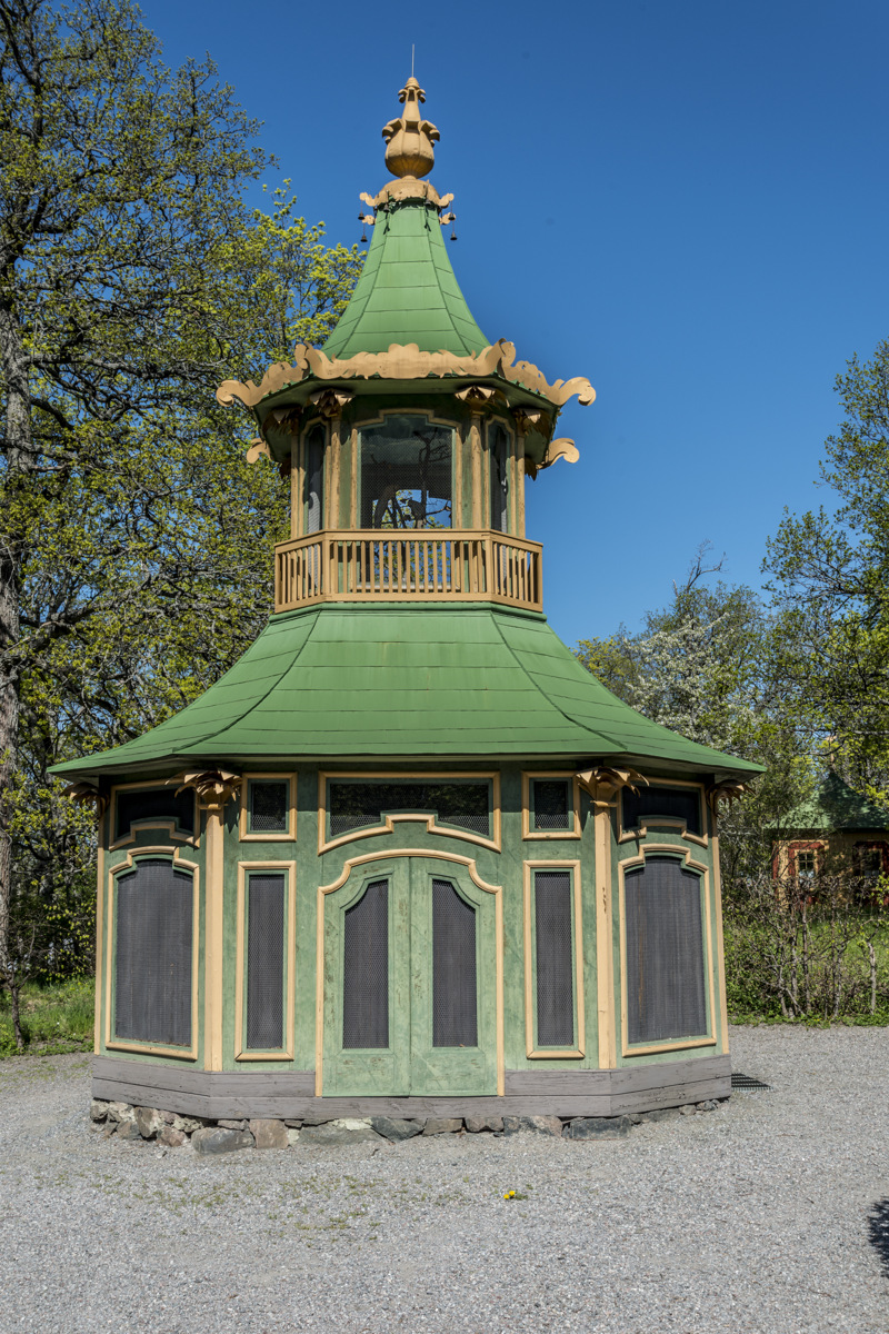 Kina slott, Drottningholm. Voljär liknande ett torn, målad i grönt, gult och svart. Den omges av häckar och stora lövträd.