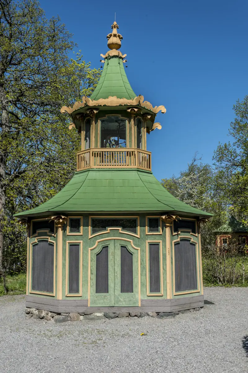 Kina slott, Drottningholm. Voljär liknande ett torn, målad i grönt, gult och svart. Den omges av häckar och stora lövträd.