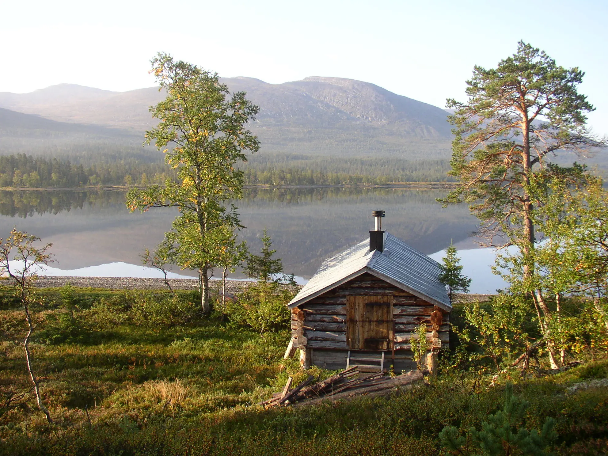 Ett litet båthus i timmer med plåt-tak invid en smal, stenig strand. Runda fjäll speglar sig i det stilla vattnet.