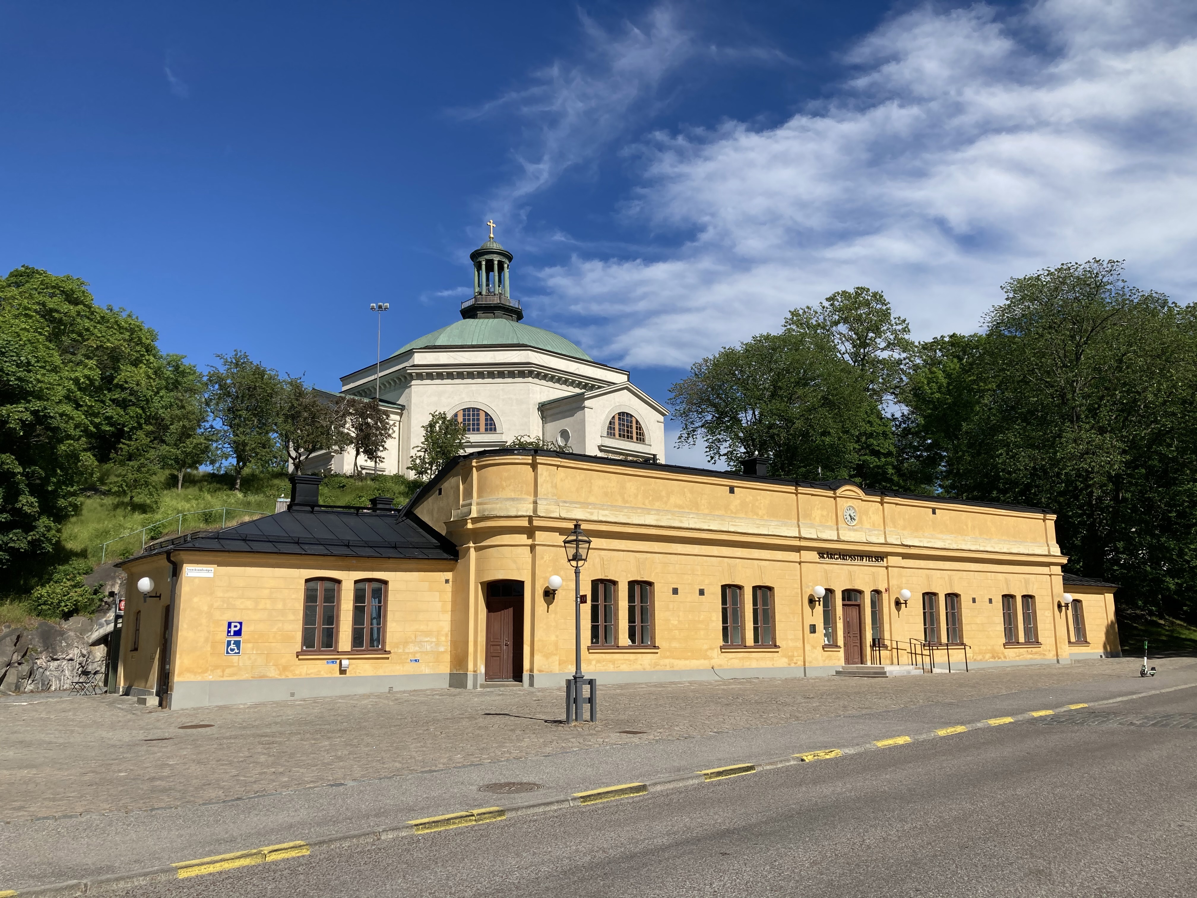 Högvakten på Skeppsholmen. Låg, gulputsad byggnad i enkel nybarock-stil. I bakgrunden syns Skeppsholmskyrkan. Sommar och grönska.