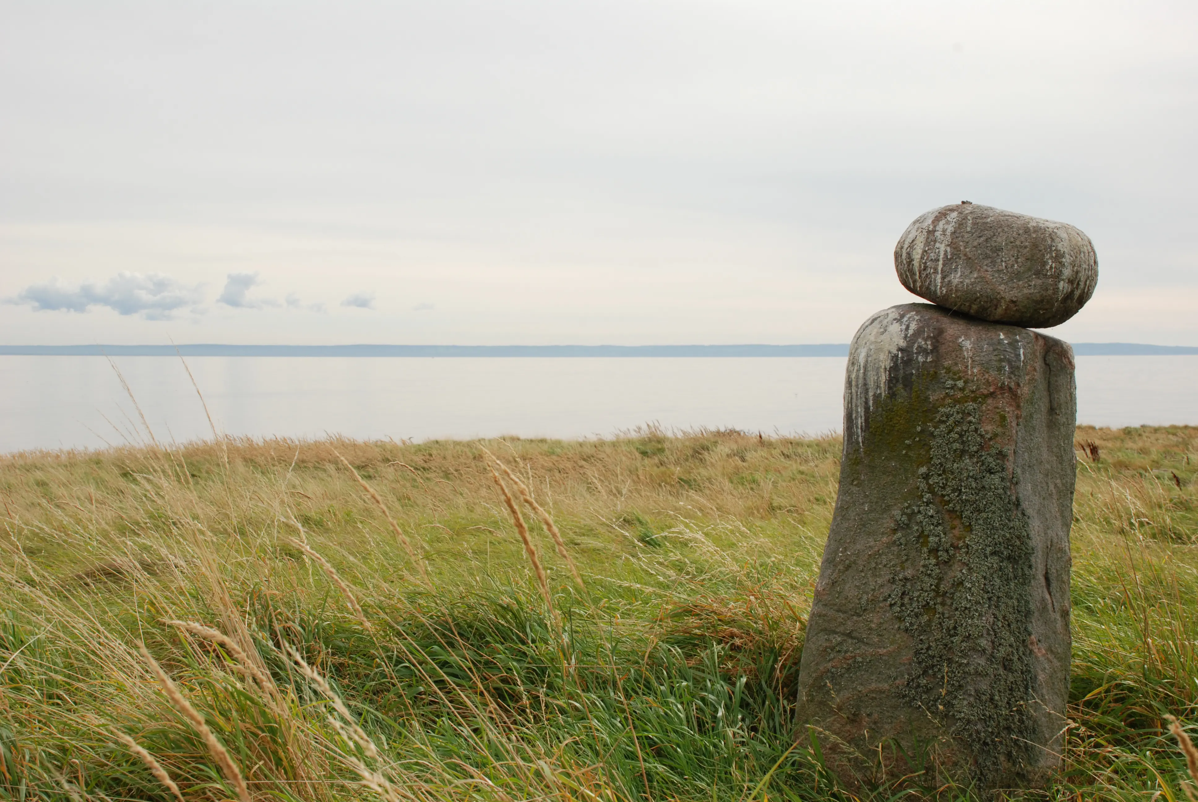 Tylön. På en gräs-beväxt kulle med utsikt över vattnet står en rest sten med en nästan rund sten ovanpå.