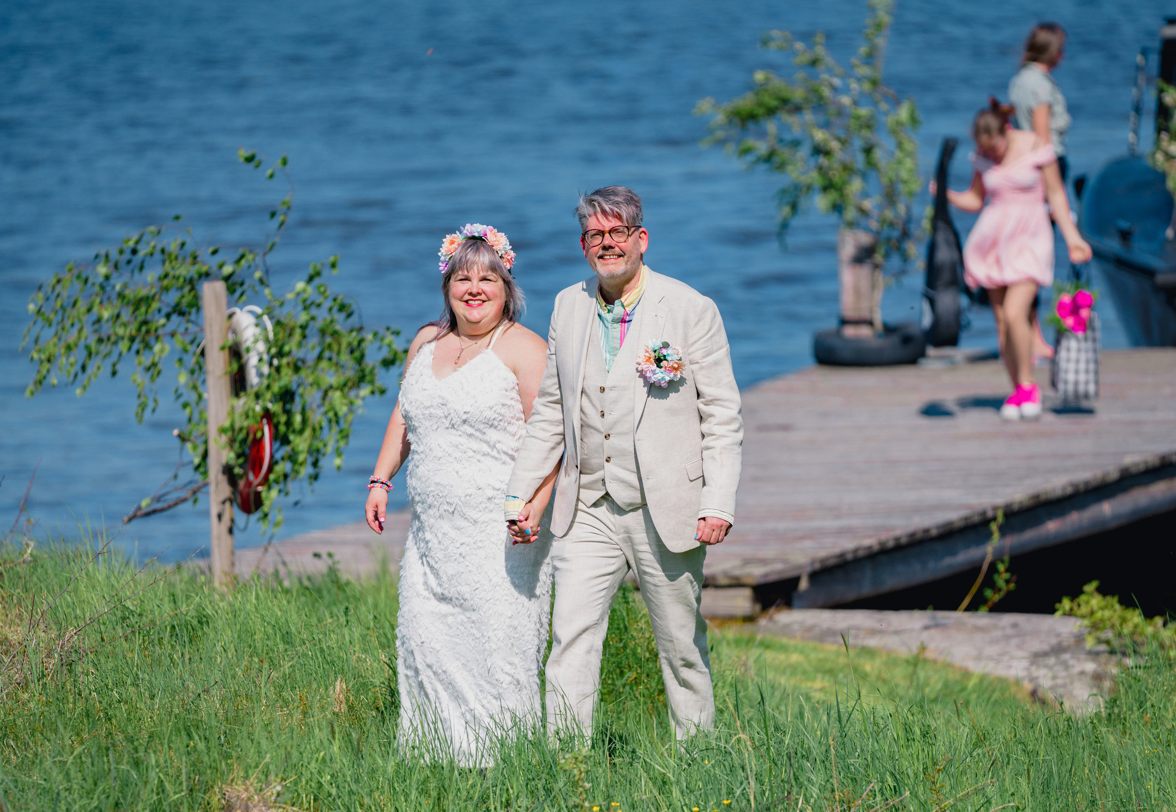 Leende brudpar i gräs med brygga och vatten i bakgrunden.