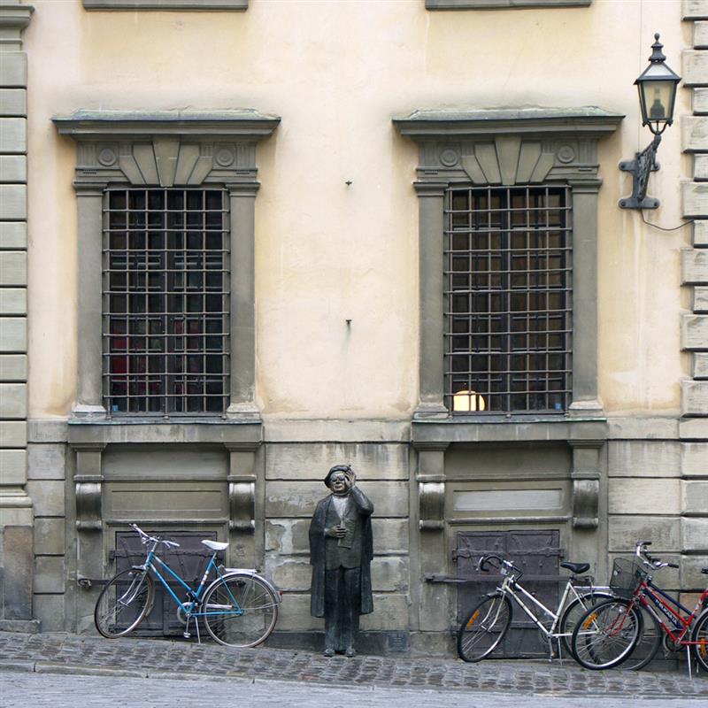 Bilden visar två fönster med galler på ett gammalt putsat hus med fönsteromfattningar . Mellan fönstren står en bronsstaty föreställande Evert Taube i basker och glasögon.