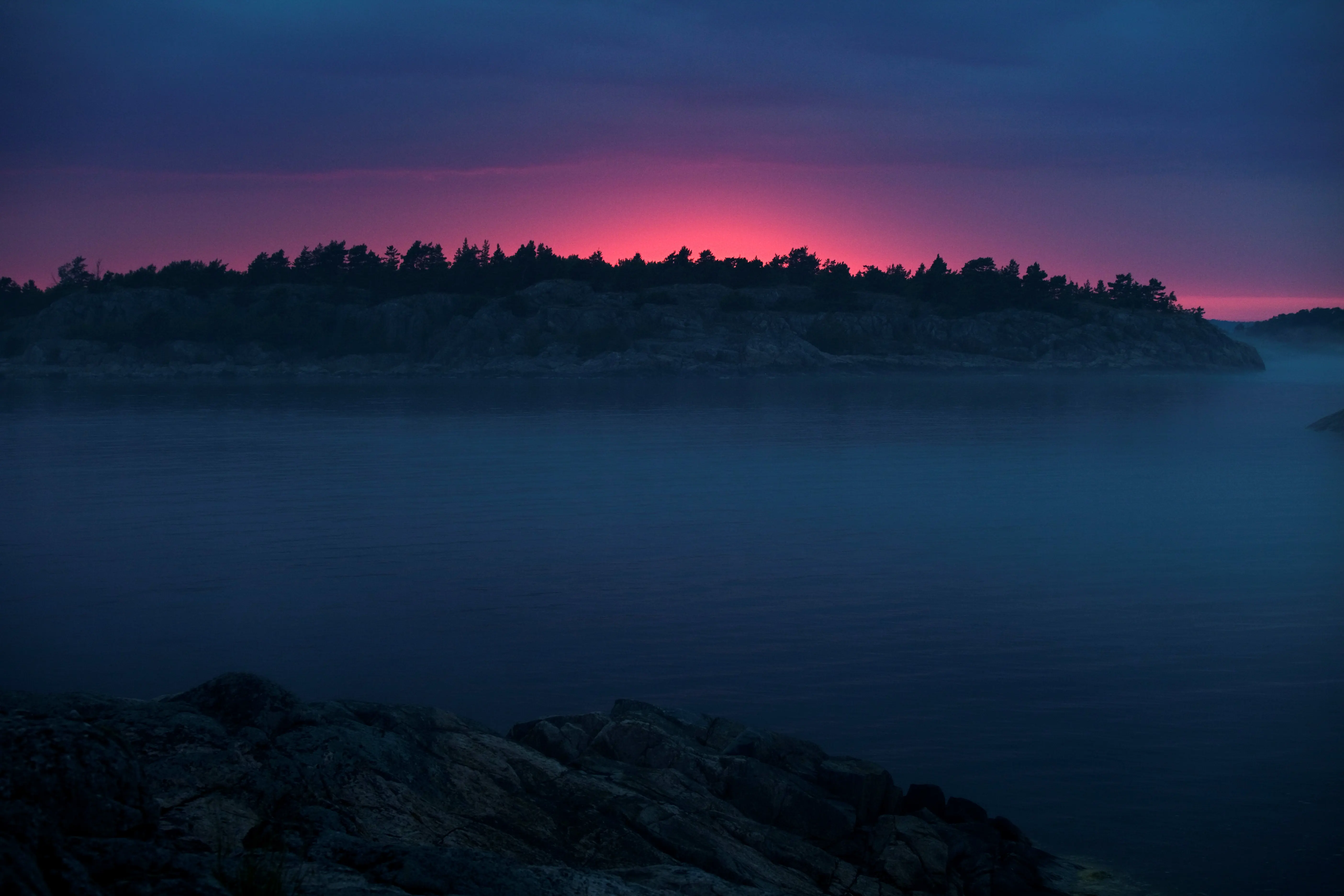 Häradsskär i skymningen. En vik täckt av dimma. Mot den röda himlen avtecknar sig träd.