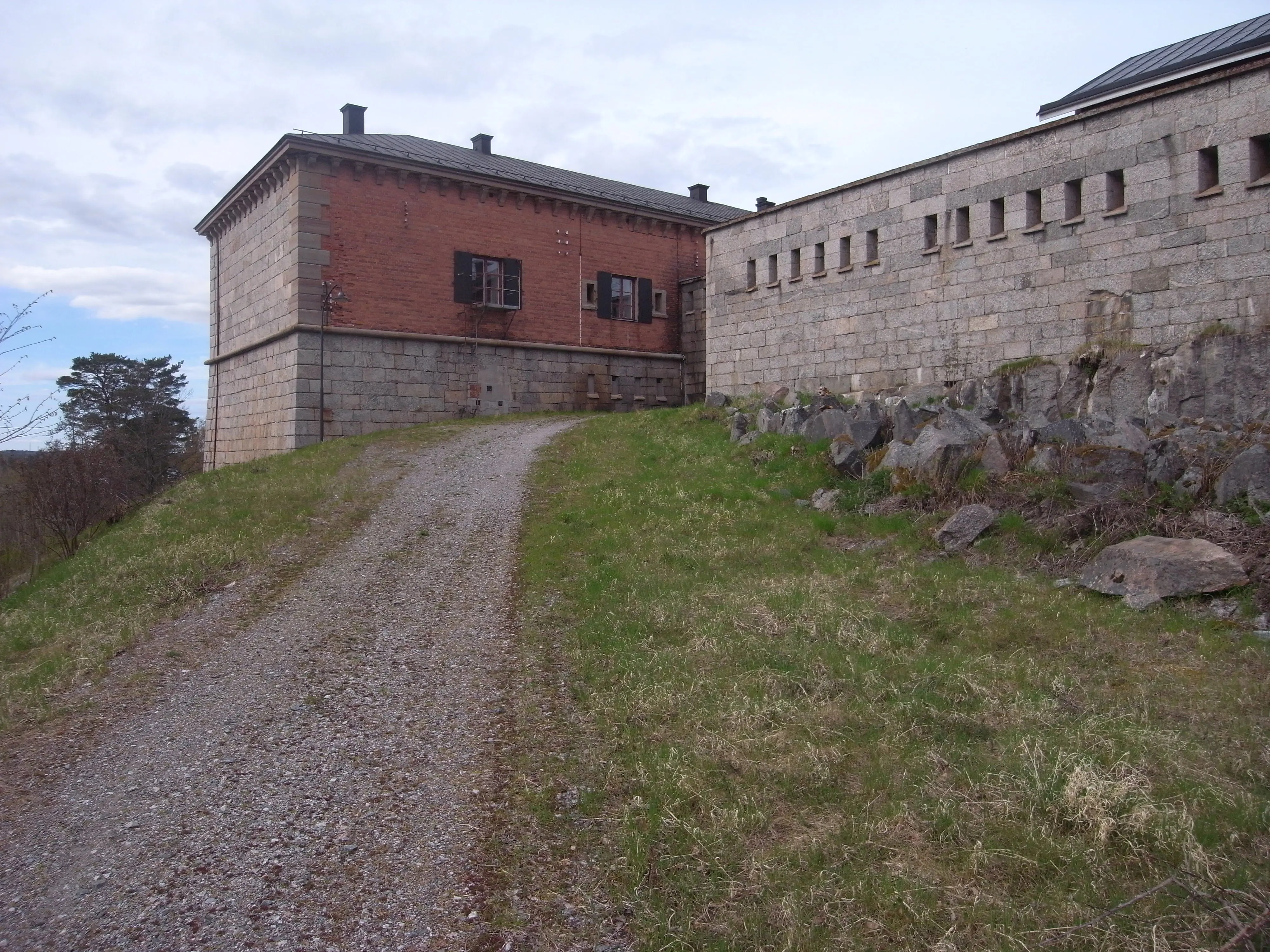 Rindö redutt. En grusväg leder upp mot en mur med små gluggar och en byggnad i sten och tegel.