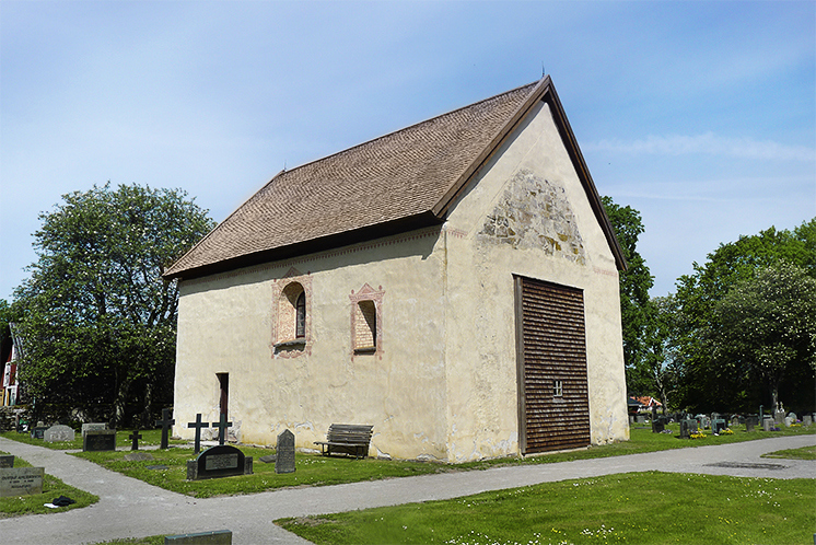 En fyrkantig liten stenkyrka med ljus fasad och brunt sluttande tak på en gräsmatta. Runt om syns en del gravstenar och kors.