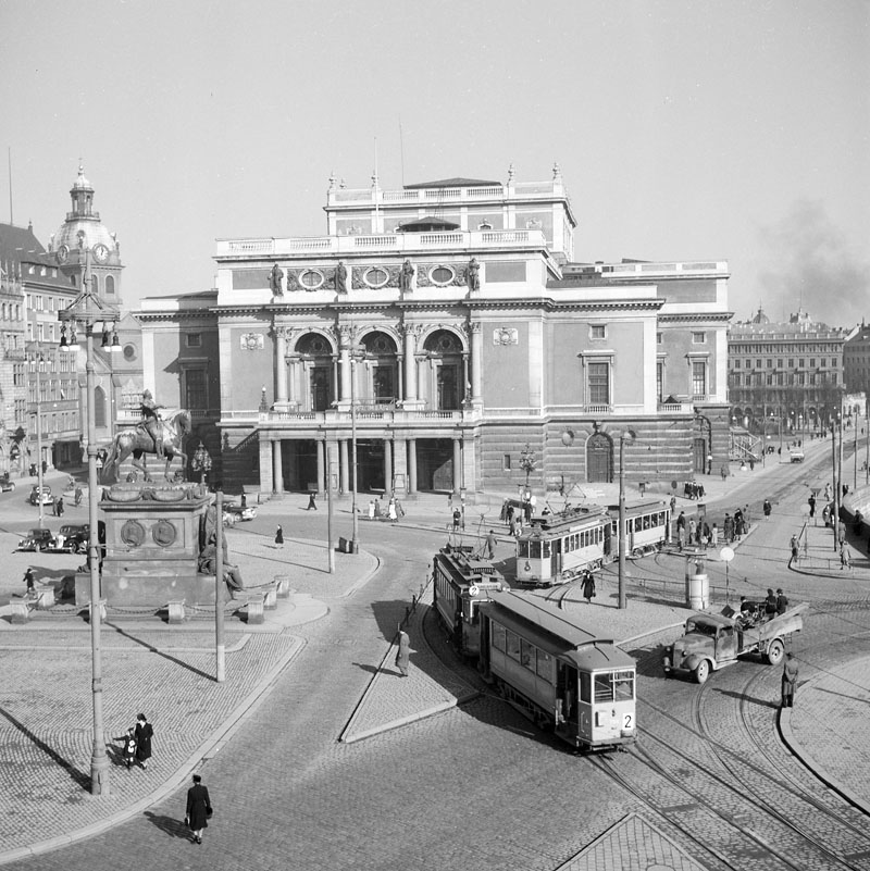 Svartvit bild av stort hus vid ett torg. På torget syns fler spårvagnar, människor och en bil.