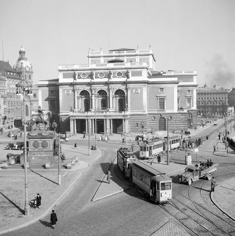 Svartvit bild av stort hus vid ett torg. På torget syns fler spårvagnar, människor och en bil.