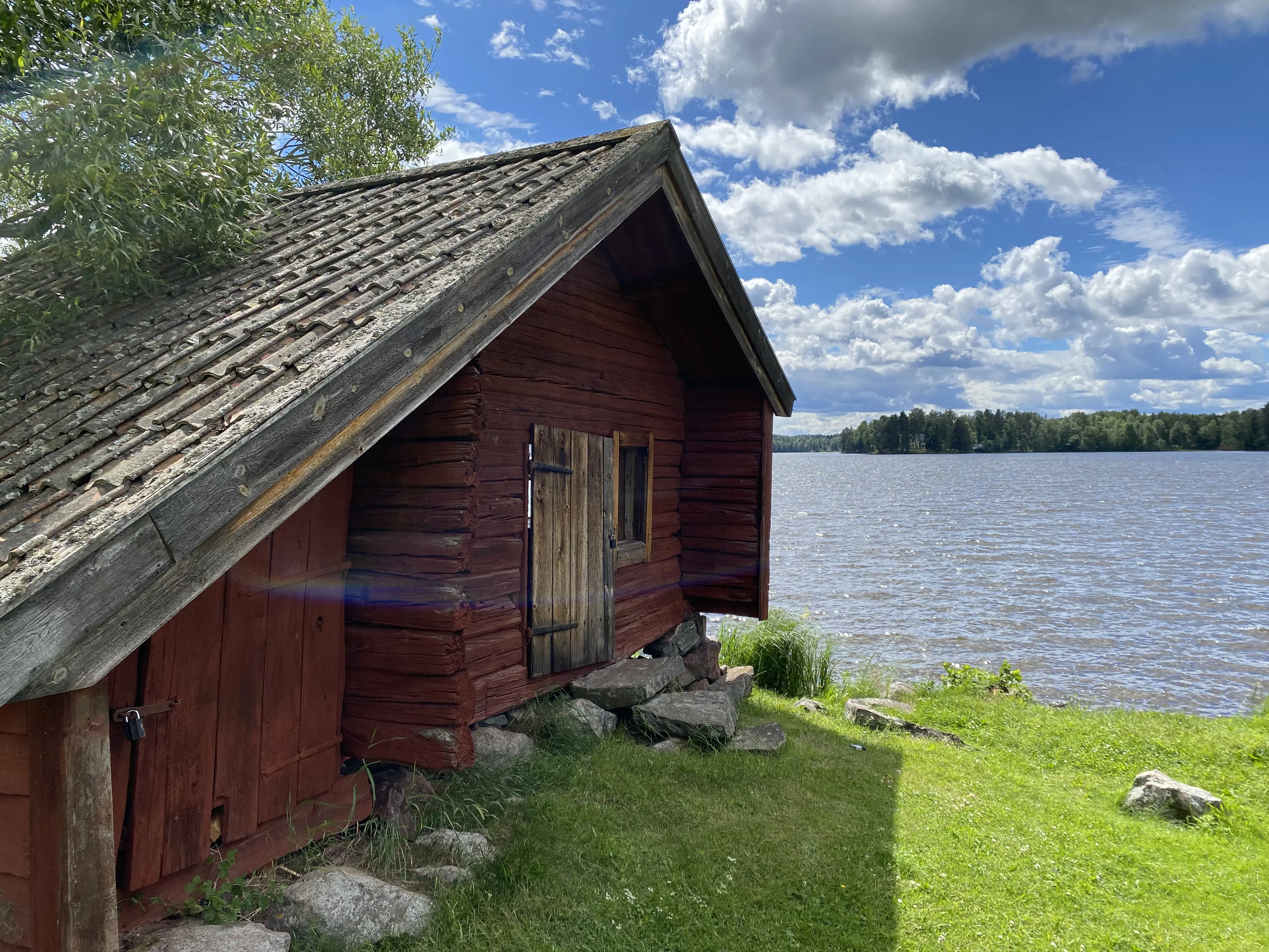 Ornässtugan. Timrat uthus vid vattnet. Sommar och grönska.