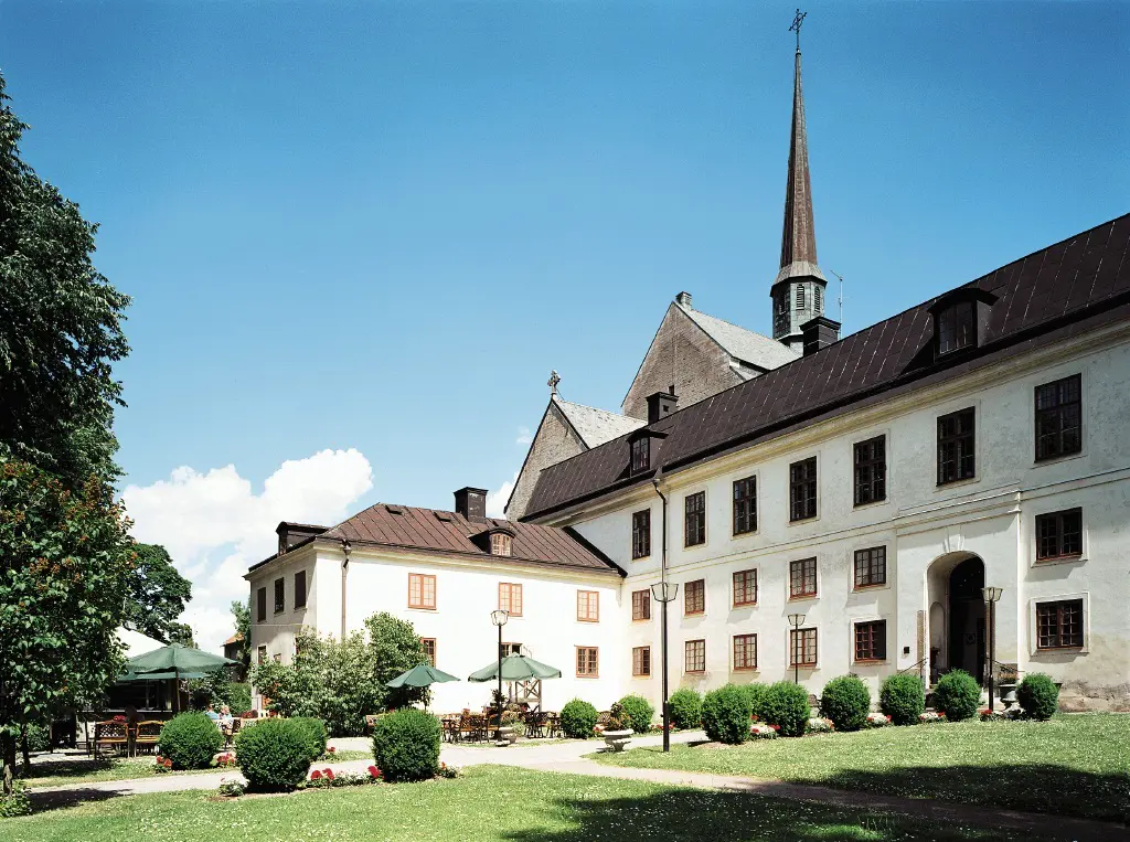 Munk-klostret i Vadstena. En vit trevåningsbyggnad, med hög entré-portal och en lägre flygel. Sommar och grönska.