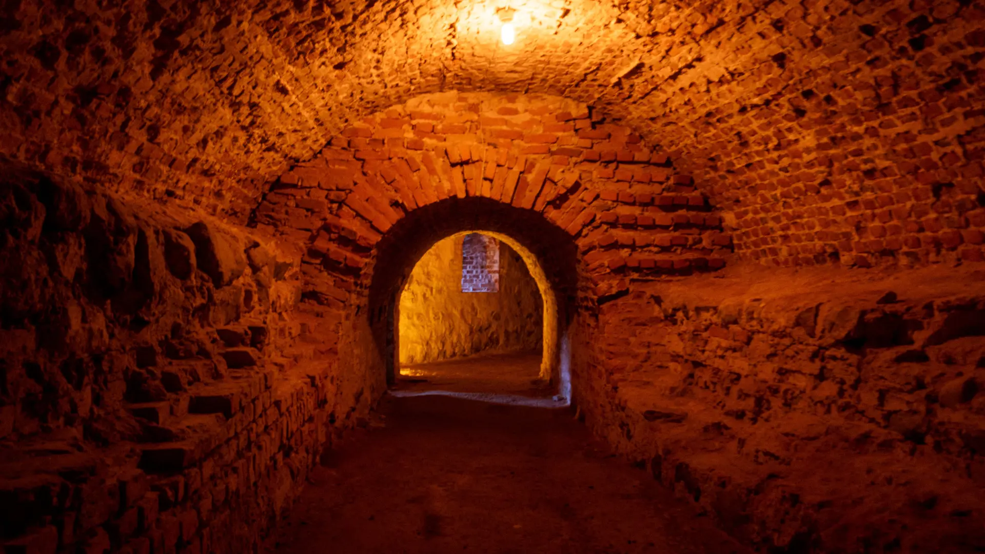 Underjordiskt valv upplyst av ett varmt gulrött ljus. i mitten syns en gång till nästa rum