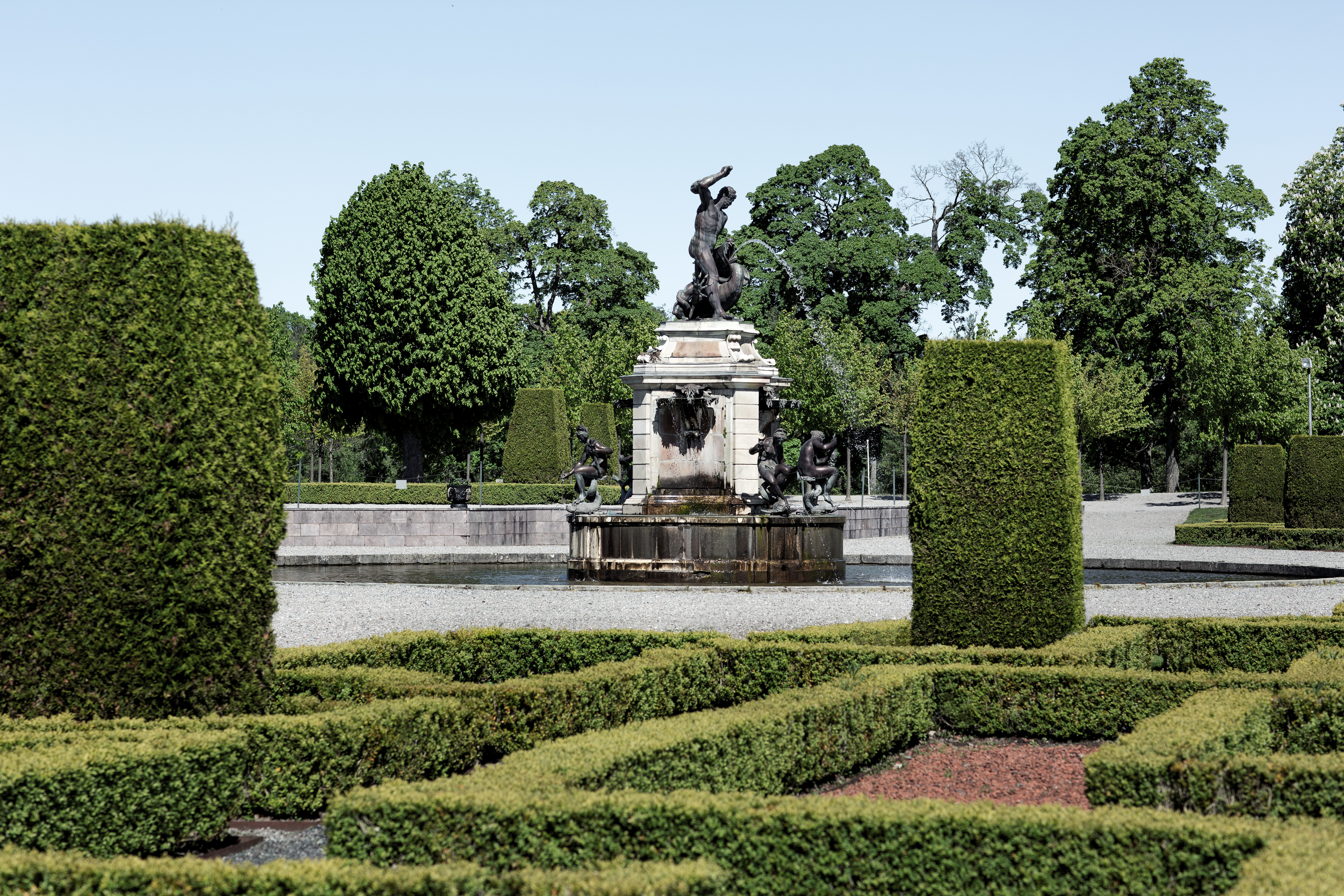 Drottningholms barockträdgård. Formklippta buxbom-häckar omger en fontän med skulpturer. Sommar och grönska.