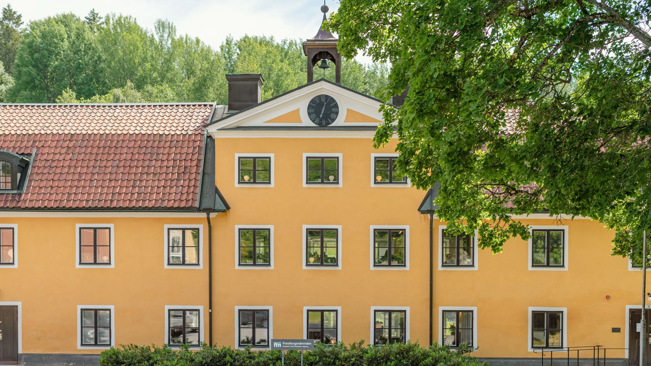 Gulputsat hus med tak av röda tegelpannor. På fasaden finns en urtavla, och längst upp på husen ett litet klocktorn.