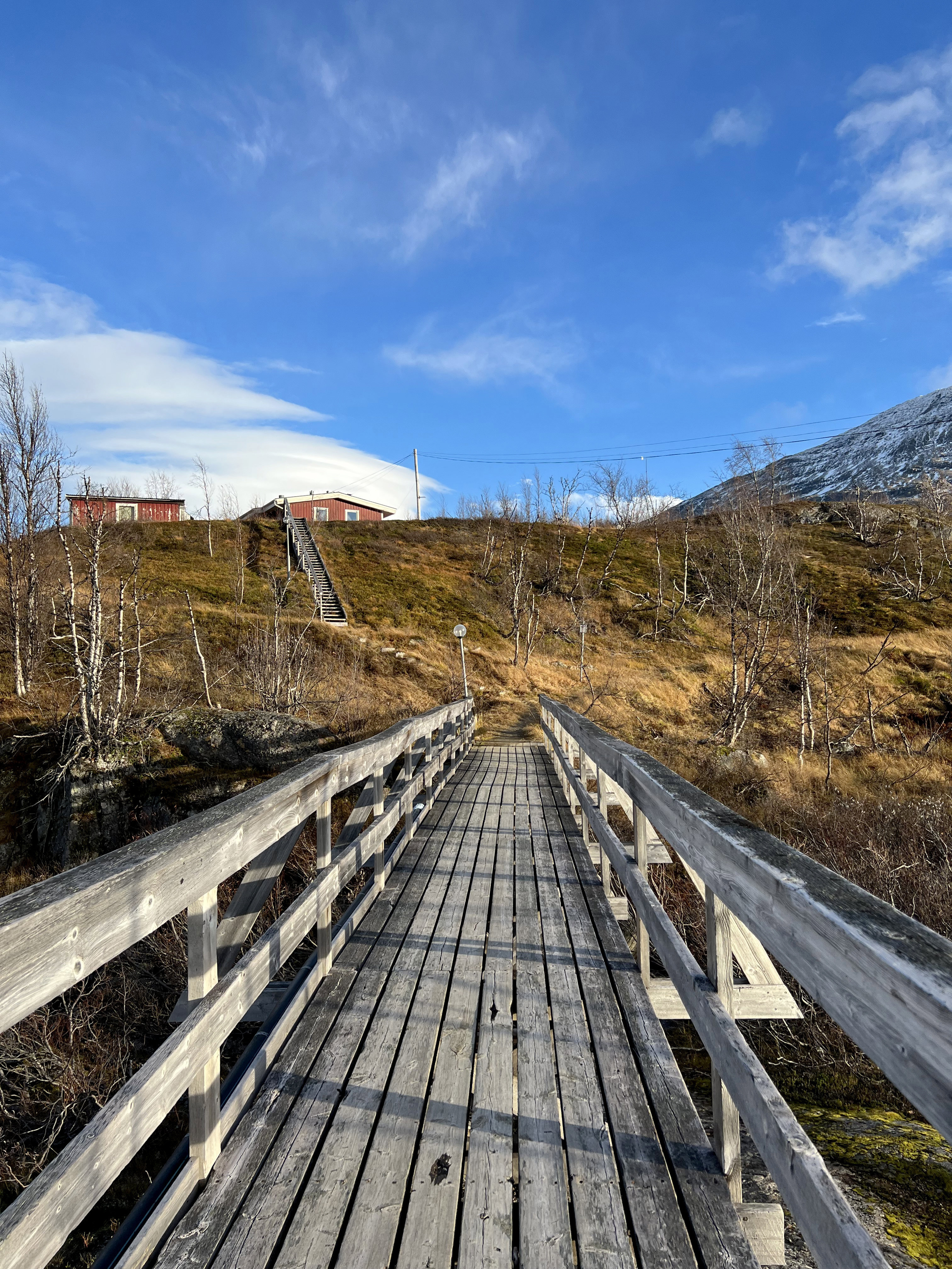Väderstationen i Katterjåkk. Två röda träbyggnader, sedda från träbron över en jokk. En trätrappa leder upp till husen, som omges av fjällbjörkar. Höst.