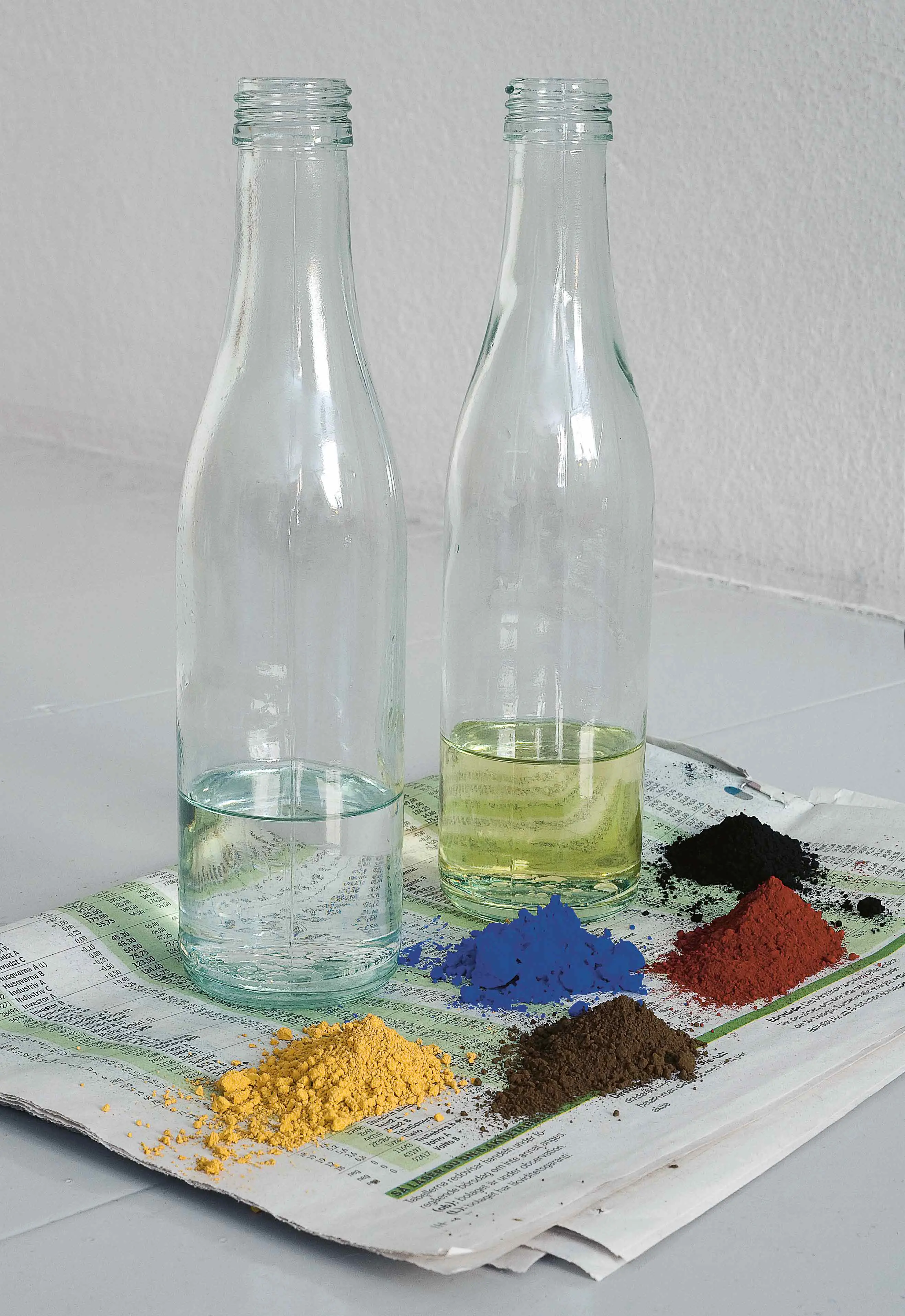 På en ihopvikt tidning står två glasflaskor delvis fyllda med vätska. Framför ligger små högar med torrpigment till olika färger.