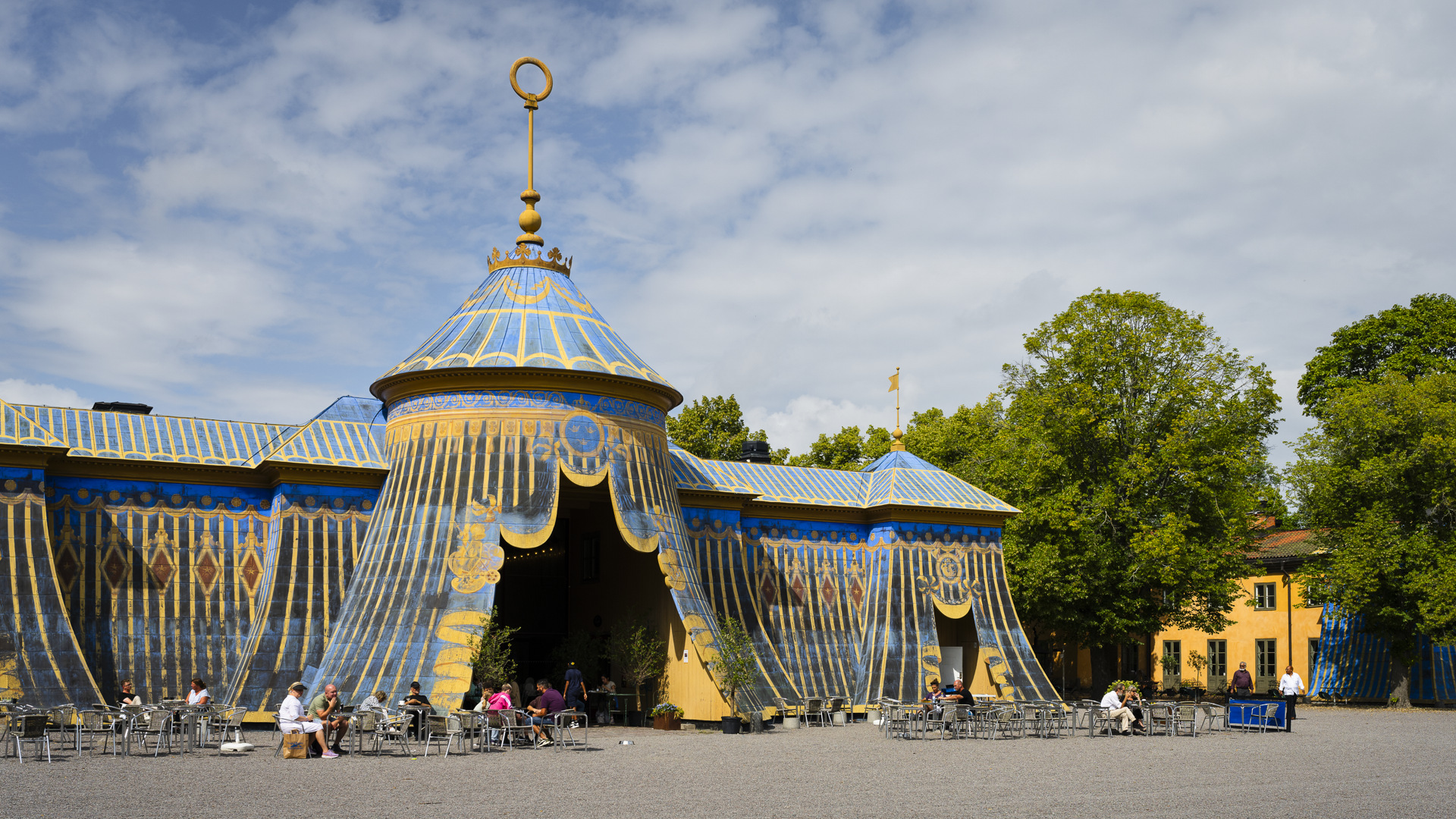 Koppartälten i Hagaparken. Plåtklädda byggnader som liknar tält, målade i blått och guldockra. På en uteservering sitter besökare.