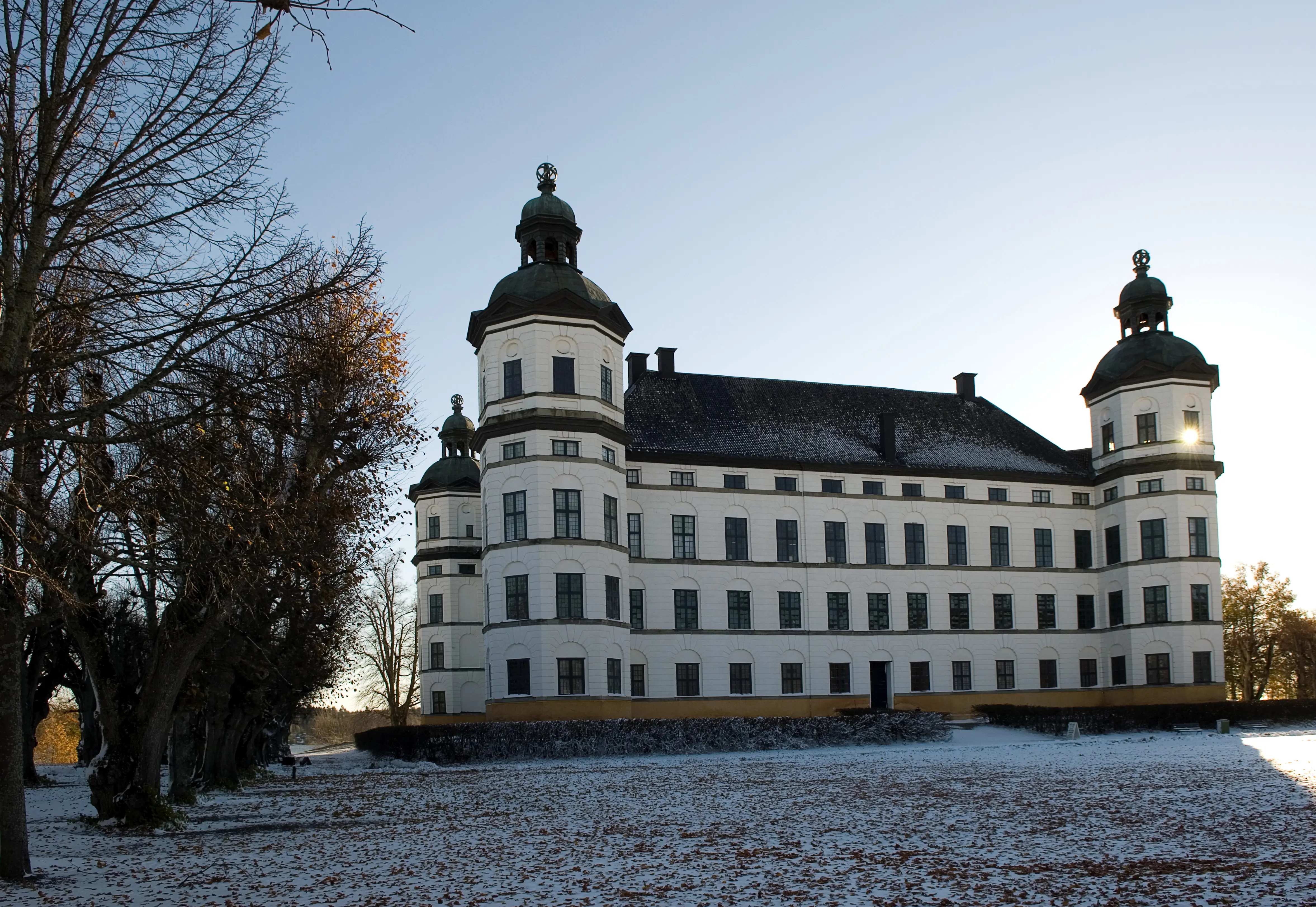 Skoklosters slott. En stor, vit byggnad med valmat tak och hörntorn. Tornen har kupoltak och lanterniner. Vinter och snö.