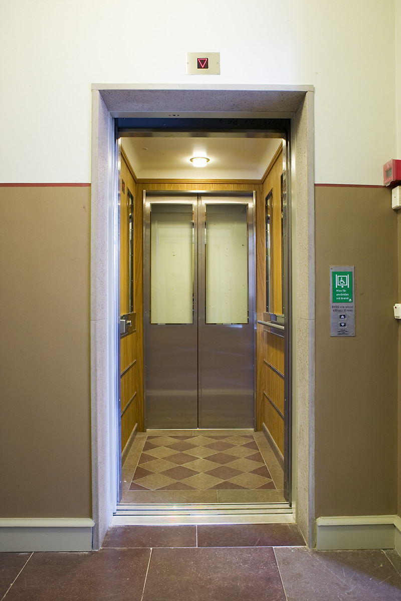 Hiss med skjutdörrar och rutigt golv. Vid knappen utanför hissen står att den får användas vid brand. 