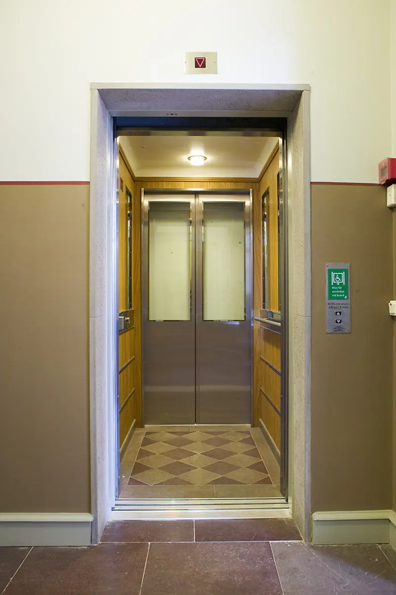 Hiss med skjutdörrar och rutigt golv. Vid knappen utanför hissen står att den får användas vid brand.