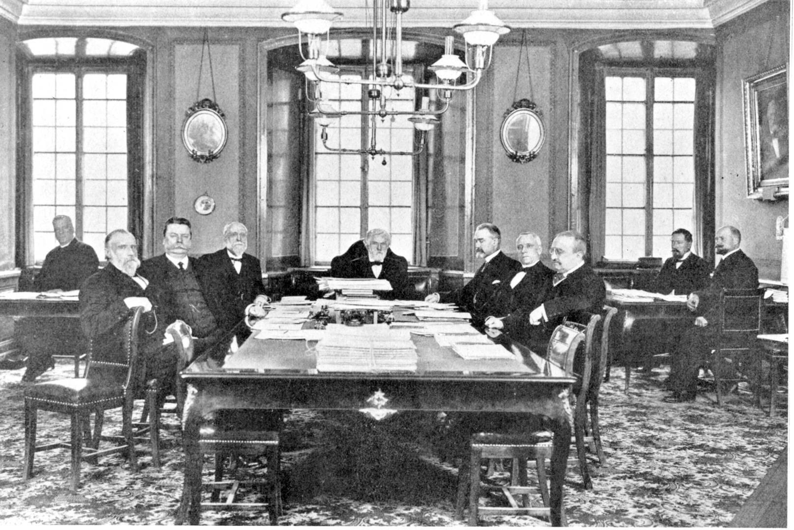 Ett gammalt svartvitt foto föreställer ett antal mörkklädda män i väst och kavaj, sekelskifte 1900. De sitter runt ett stort bord i en sal med inredning från 1700-talet.