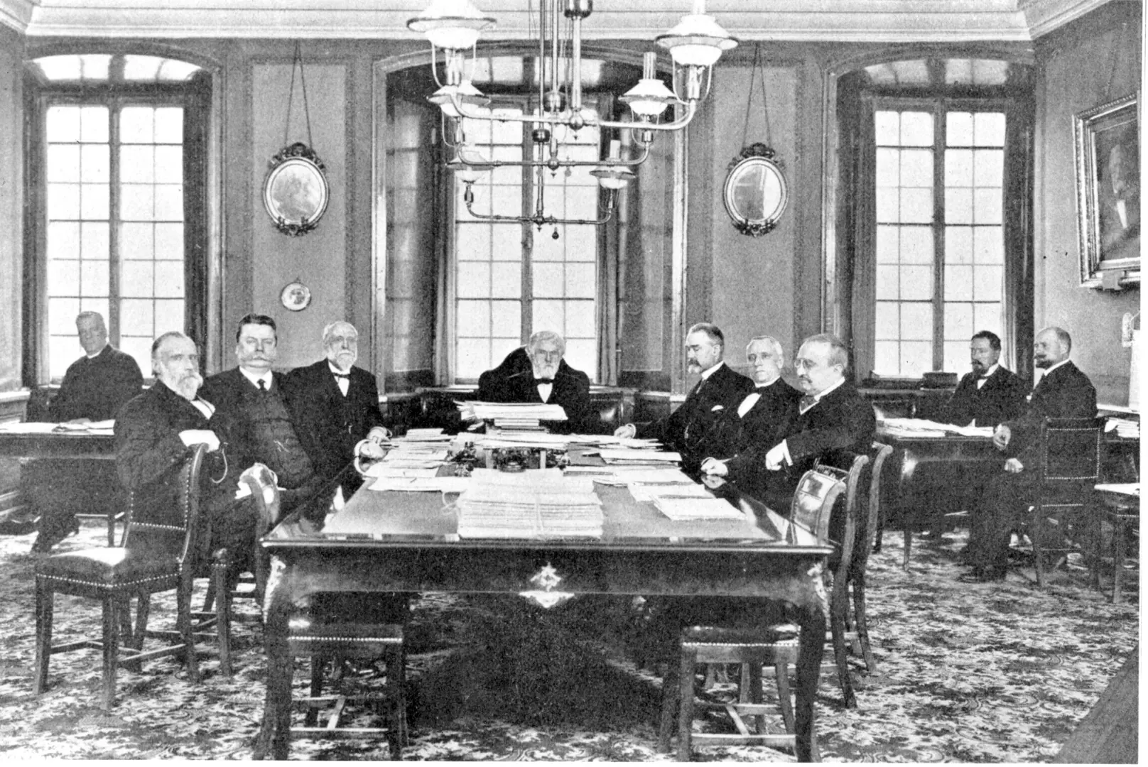 Ett gammalt svartvitt foto föreställer ett antal mörkklädda män i väst och kavaj, sekelskifte 1900. De sitter runt ett stort bord i en sal med inredning från 1700-talet.