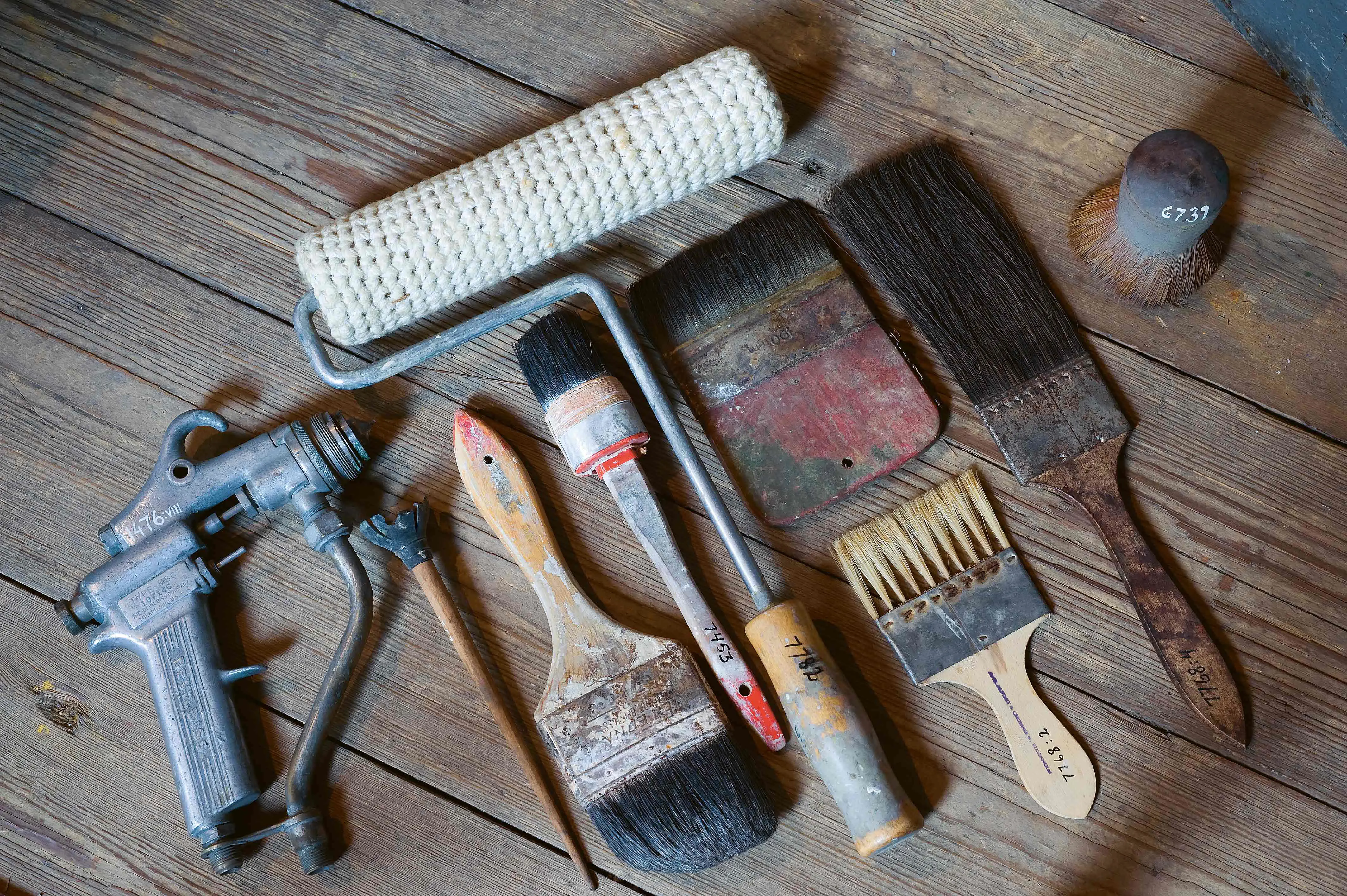 På ett trägolv ligger i rad en färgpistol, penslar och borstar i olika storlekar och utformande samt en målarroller.