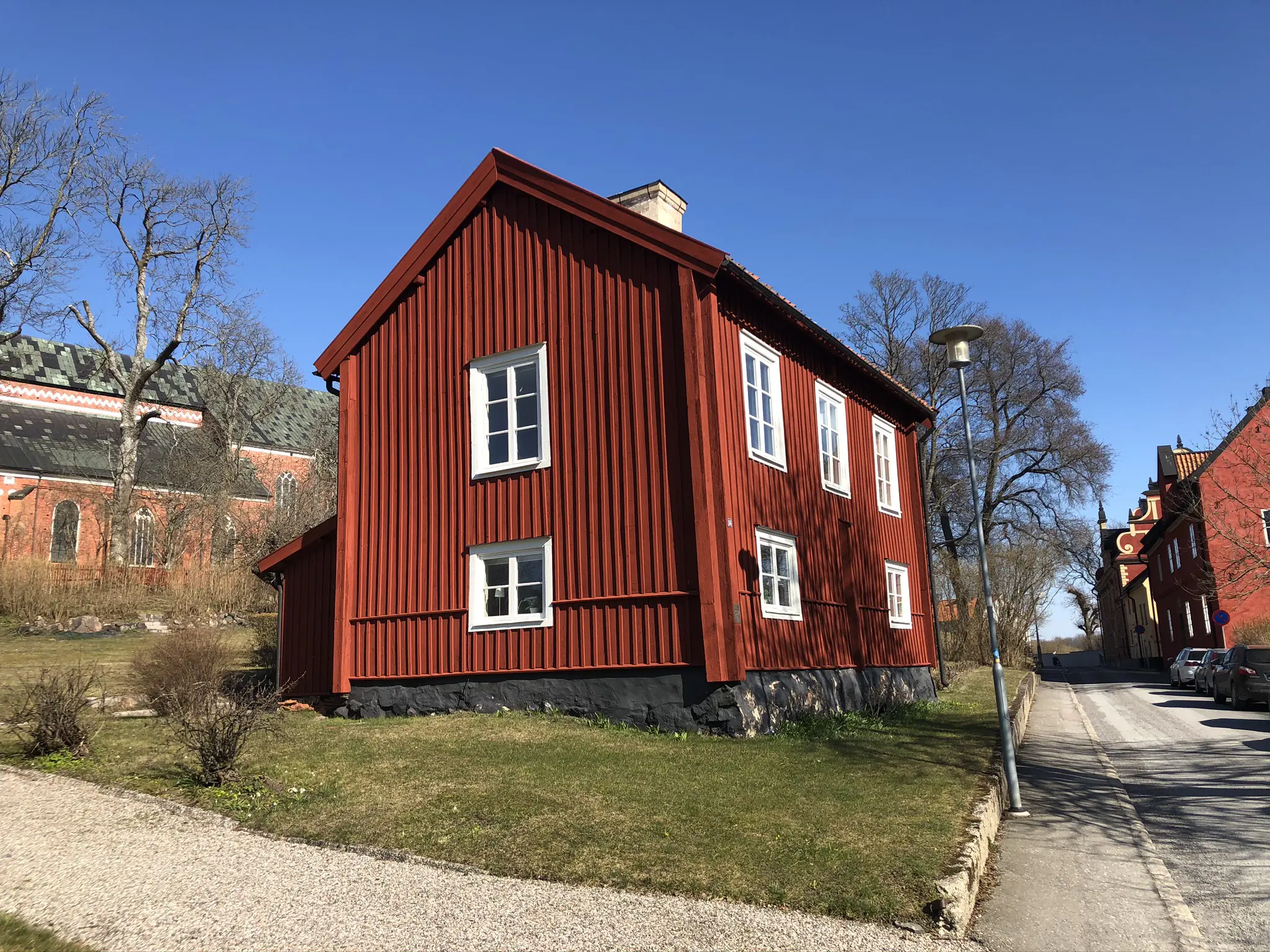 Lycktan, ett rött trähus med vita fönsterbågar, sett från gatan, med domkyrkan i bakgrunden. Tidig vår.