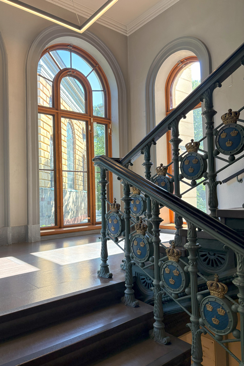 Armémuseum. Trappavsats med stengolv och höga, rund-bågiga fönster. Ett gjutjärns-räcke med tre kronor går längs trappan.