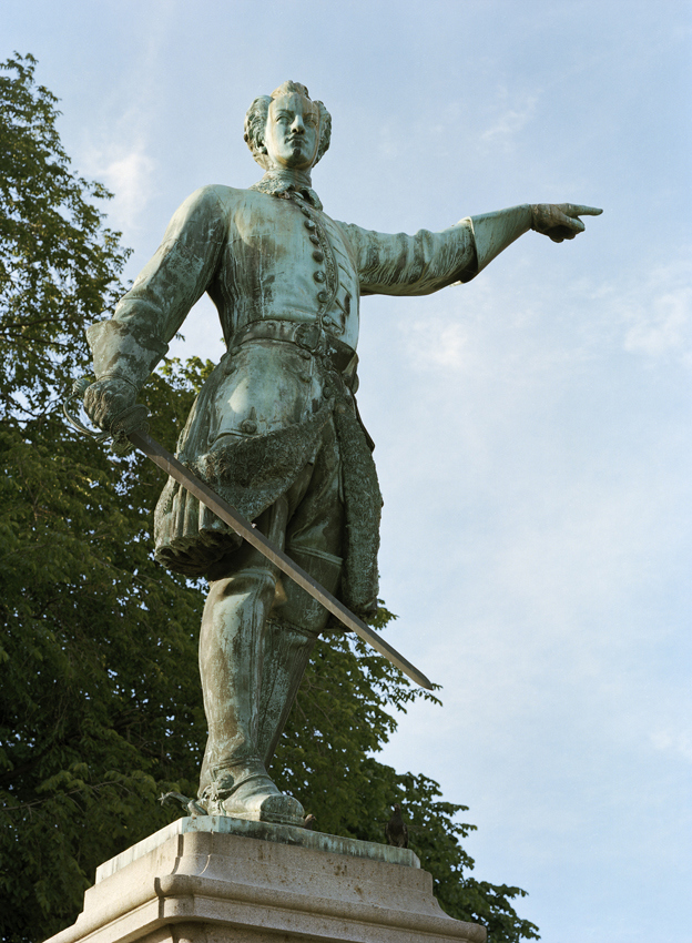 Karl den tolftes staty i Kungsträdgården. Ärgad brons-staty av en man som pekar åt sidan med vänstra handen. I den högra håller han ett svärd.