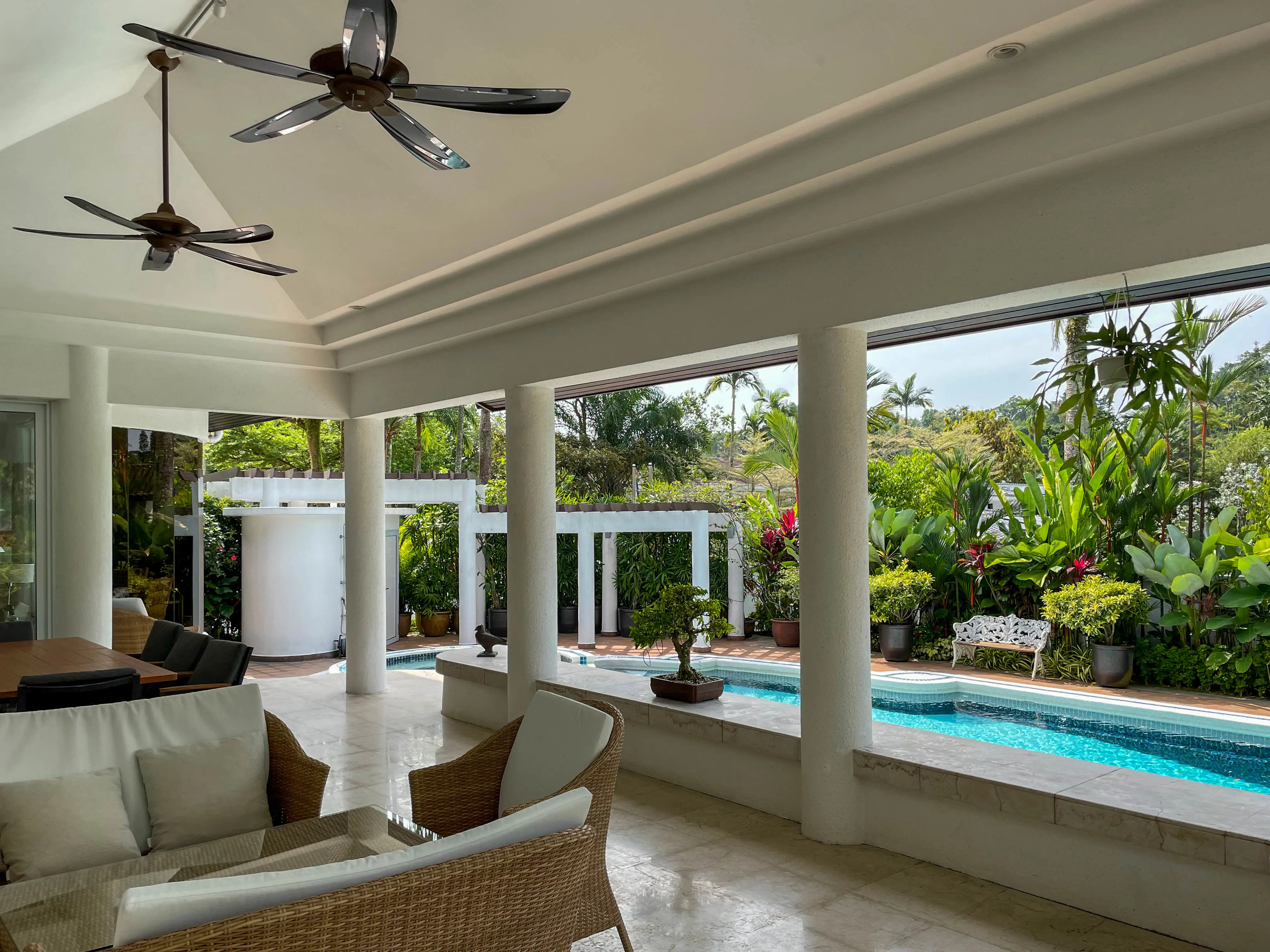 En utaplats med pelare och fläktar i taket. Utemöbler i rotting. En pool utanför.