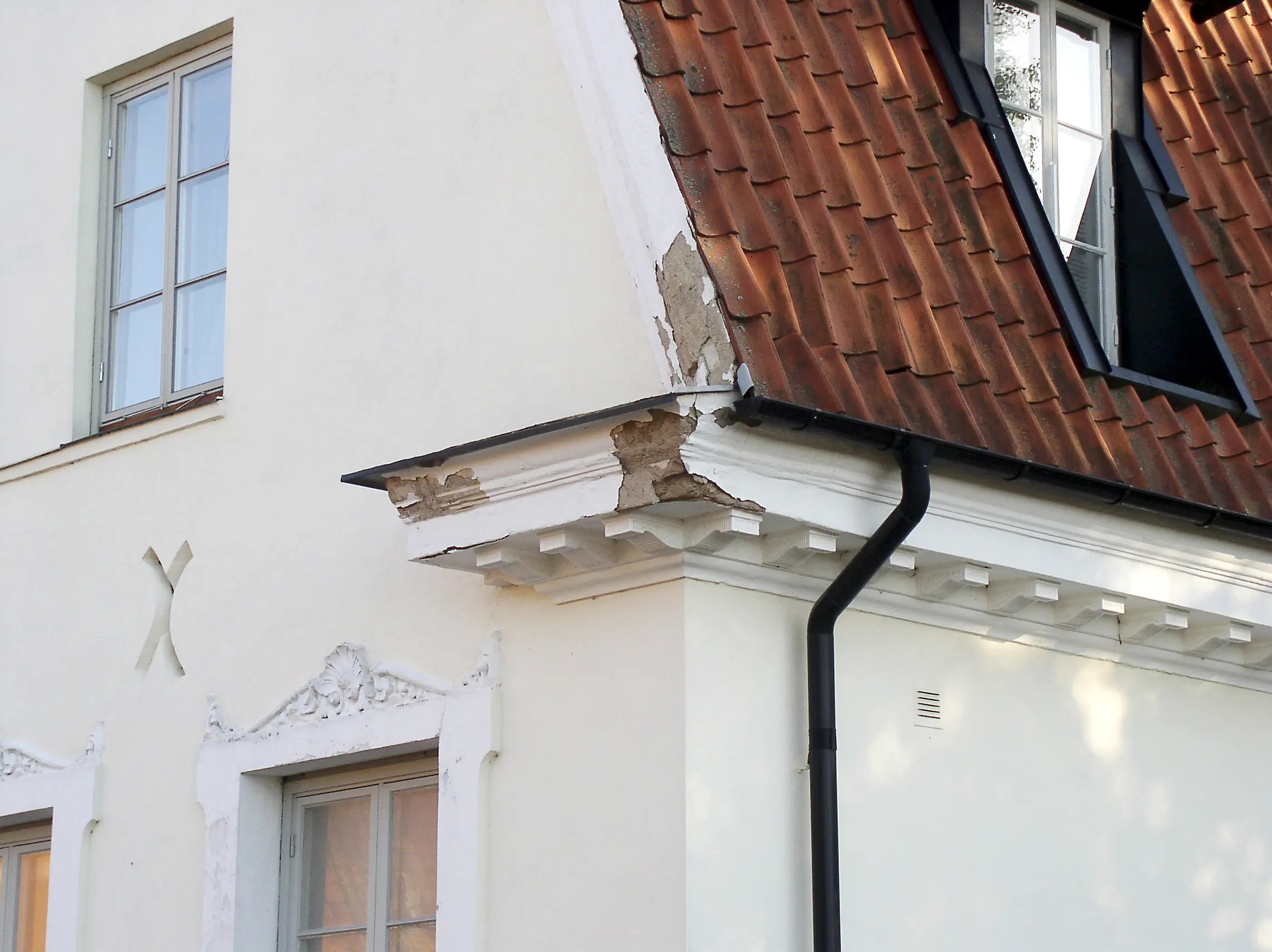 En husgavel med vit putsad fasad, ett rött tegeltak. Fasaden har skador i sig, stora bitar av den vita färgen som fallit bort.