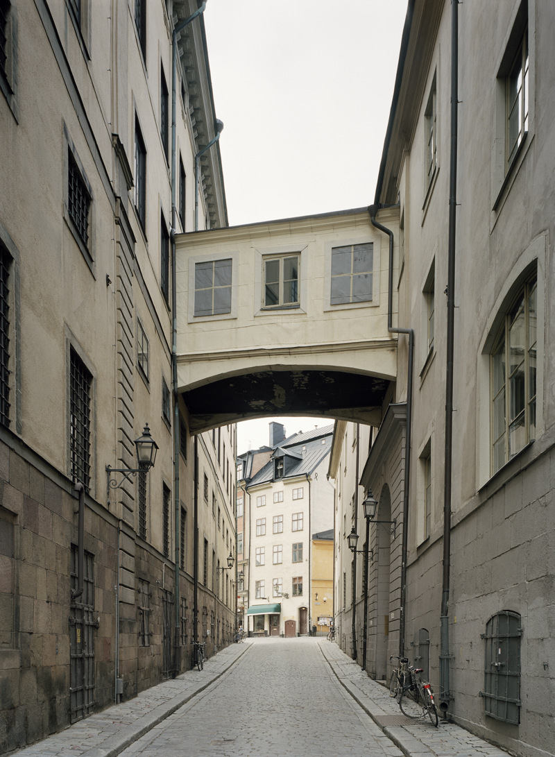 Bilden visar Norra Bankogränd i Gamla Stan Stockholm. Gränden är stenlagd och fasaderna är putsade i gulvitt. Två våningar upp syns en inbyggd gångbro som bilder samman två byggnader.