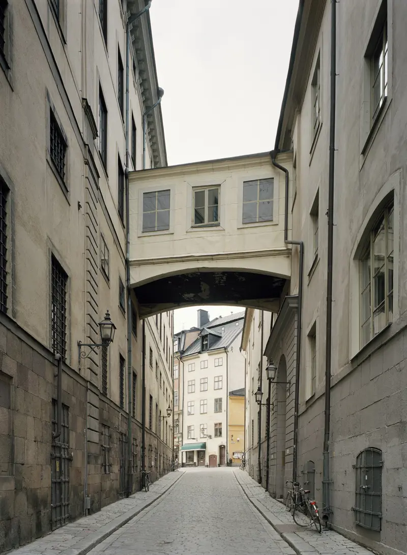 Bilden visar Norra Bankogränd i Gamla Stan Stockholm. Gränden är stenlagd och fasaderna är putsade i gulvitt. Två våningar upp syns en inbyggd gångbro som bilder samman två byggnader.
